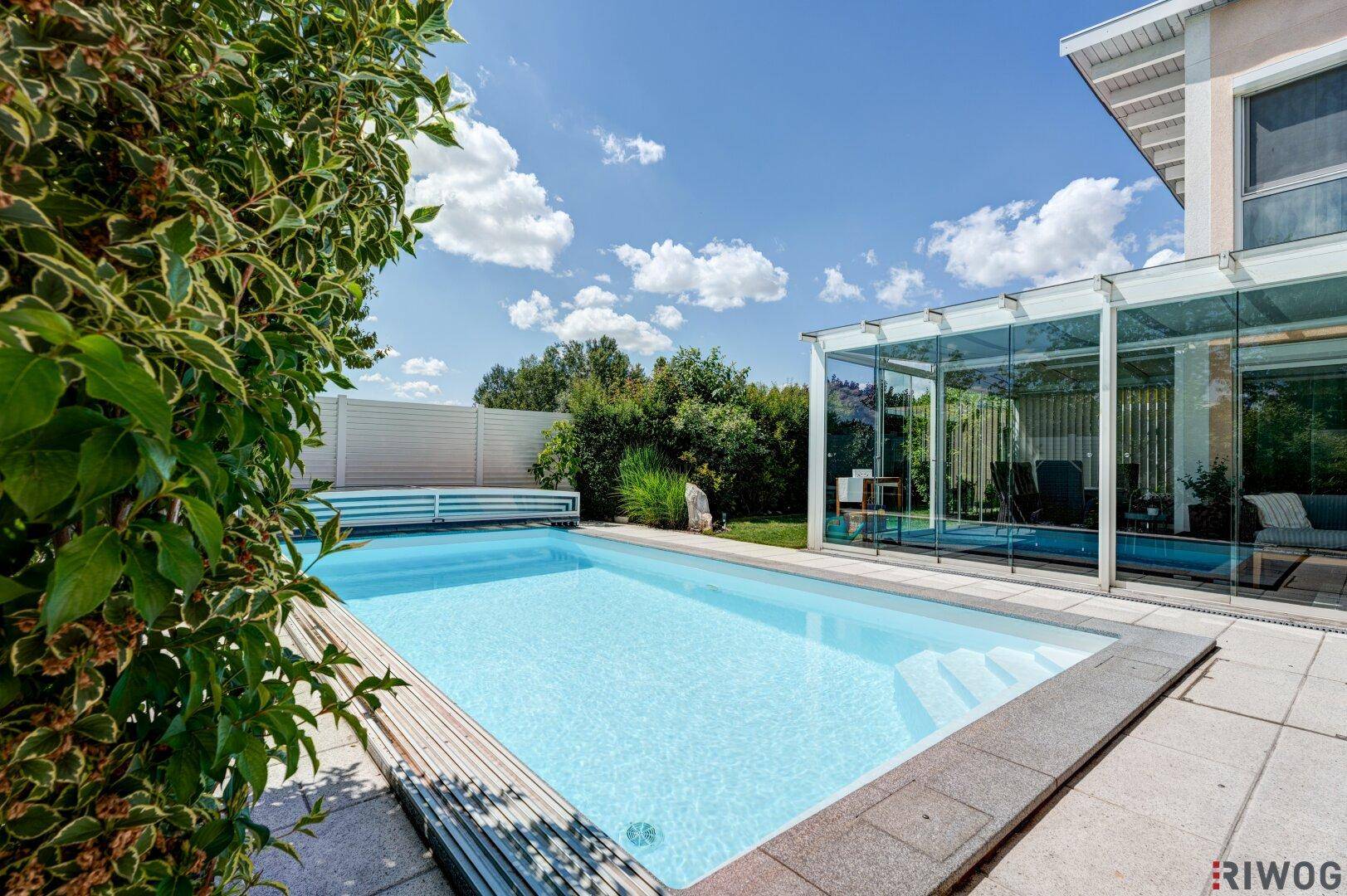 Blauer Swimmingpool umgeben von üppigem Grün und einer modernen Hausfassade.