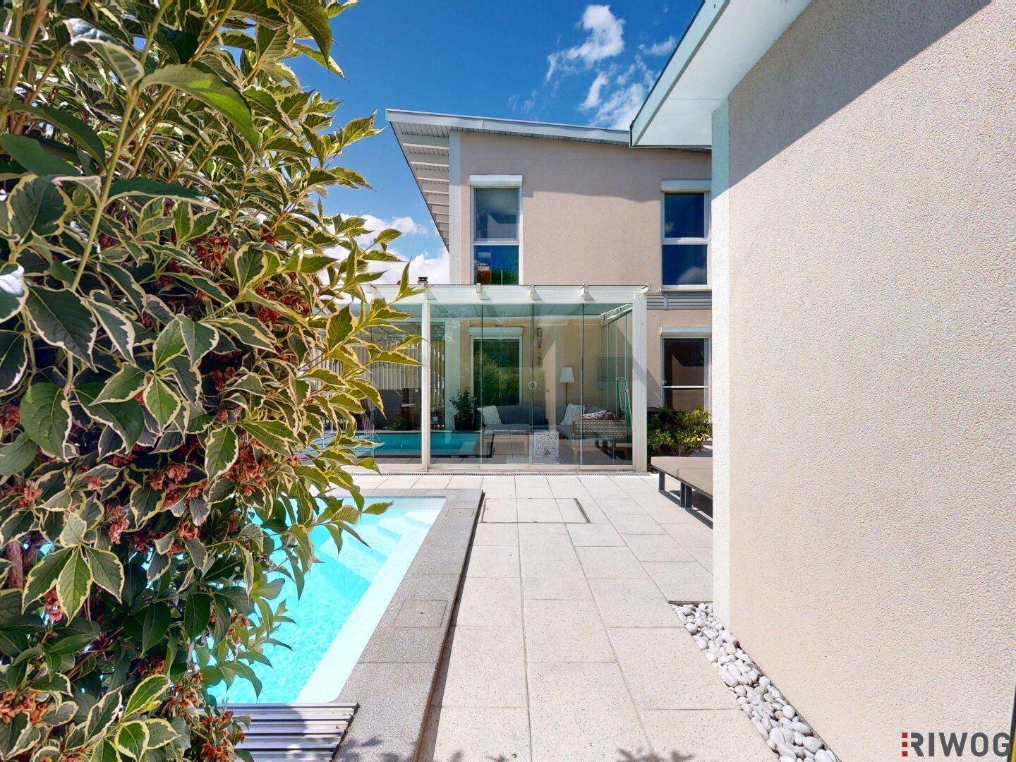 Moderne Hausfassade mit Poolbereich und verglastem Wintergarten unter blauem Himmel.