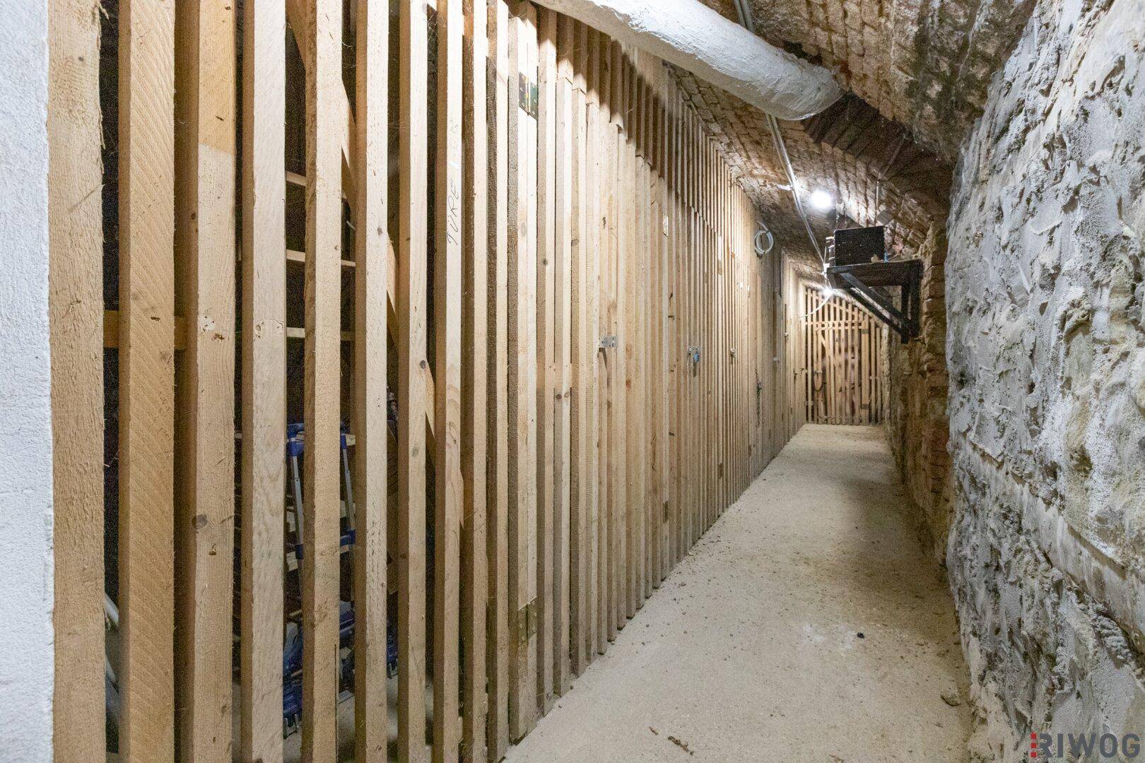 Langer Kellergang mit Holzverschlägen und unverputzten Steinwänden, ideal als Lagerraum.