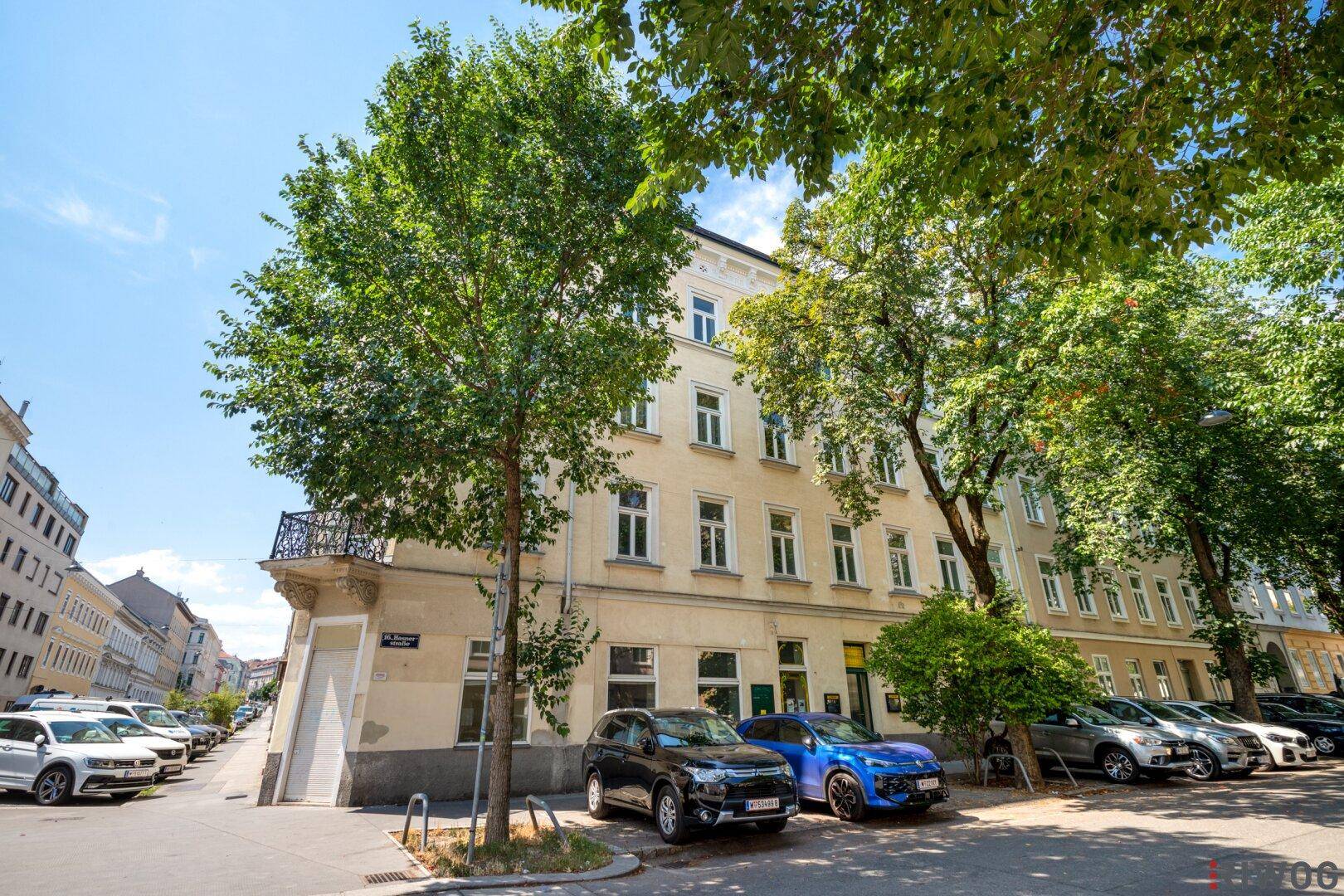 Gebäudeansicht an einem sonnigen Tag mit Bäumen und geparkten Fahrzeugen auf der Straße.