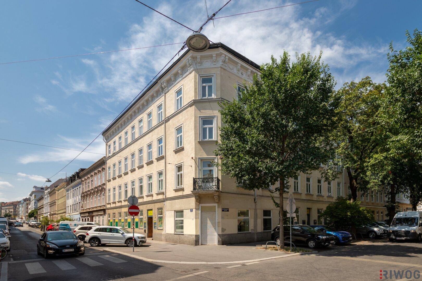 Gebäudeansicht am Tag mit geparkten Autos und Bäumen an der Straßenecke.