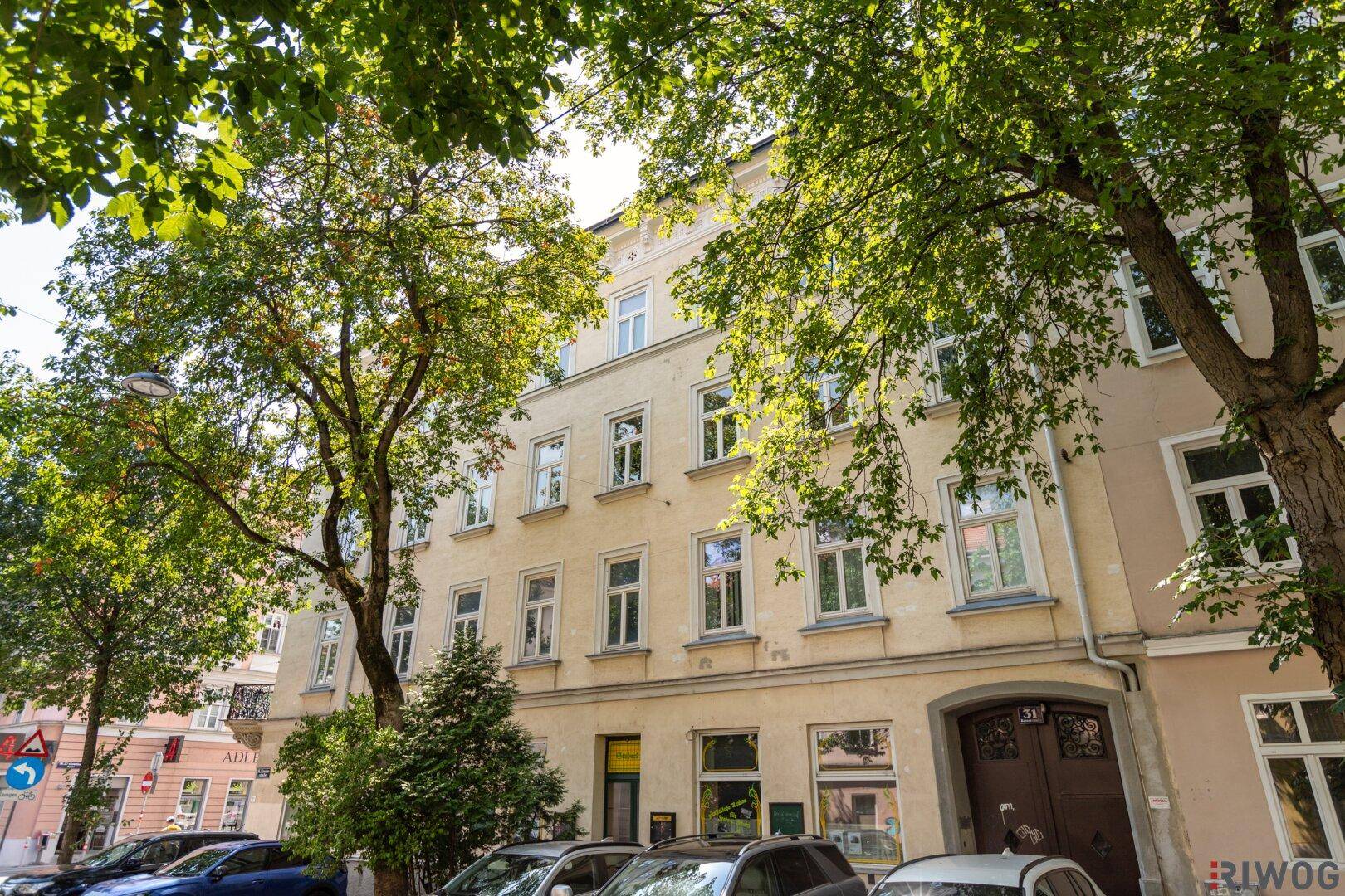 Fassade eines Altbaus mit vielen Fenstern und Bäumen im Vordergrund an einem sonnigen Tag.
