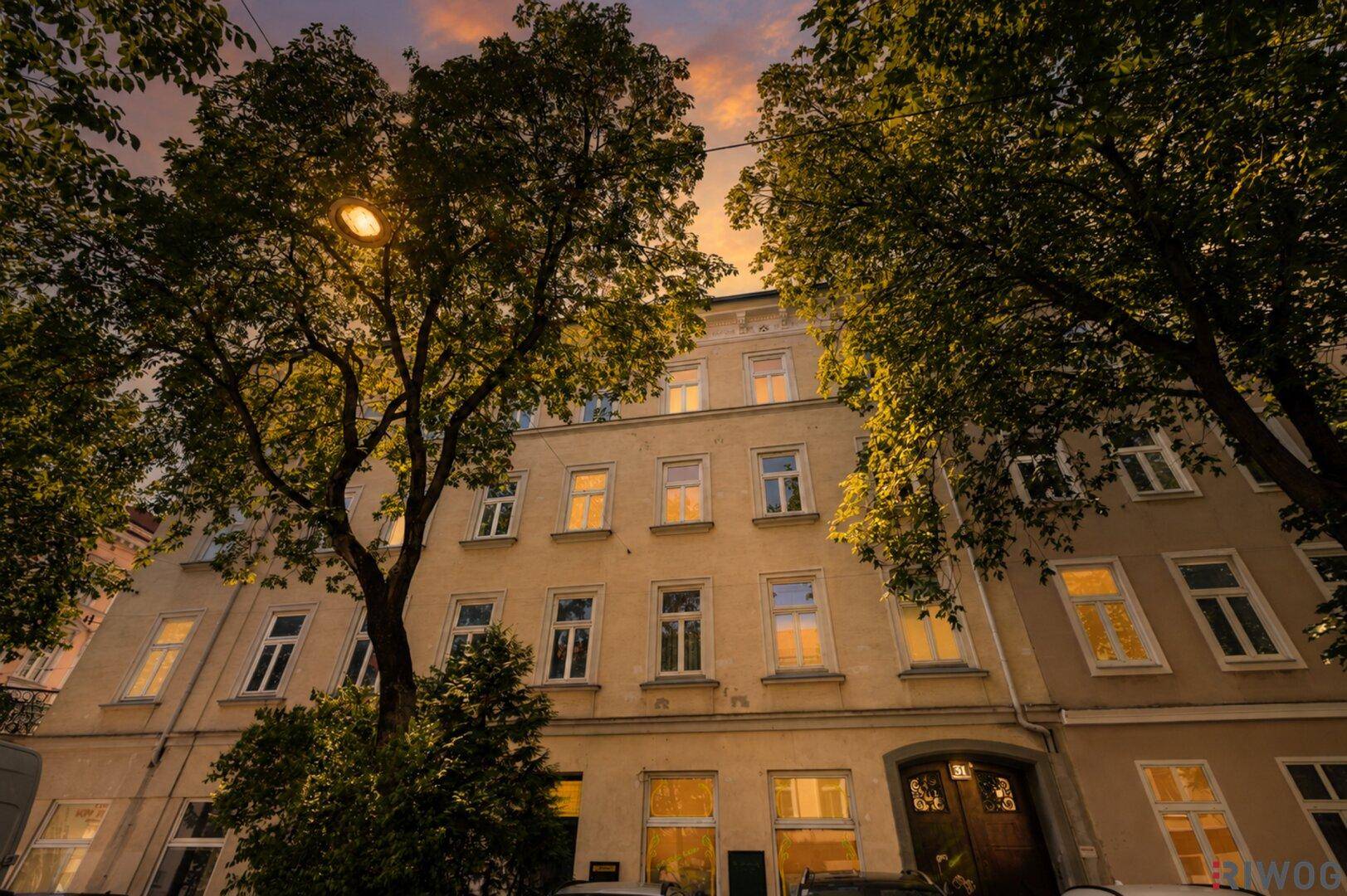 Gebäudefassade bei Sonnenuntergang mit beleuchteten Fenstern und Bäumen im Vordergrund.