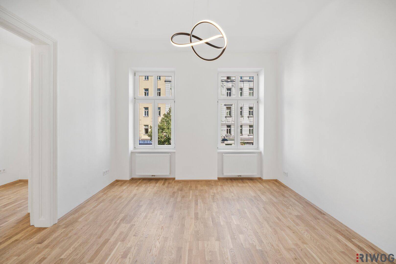 Helles, leeres Zimmer mit Parkettboden und zwei großen Fenstern, die viel Tageslicht hereinlassen.