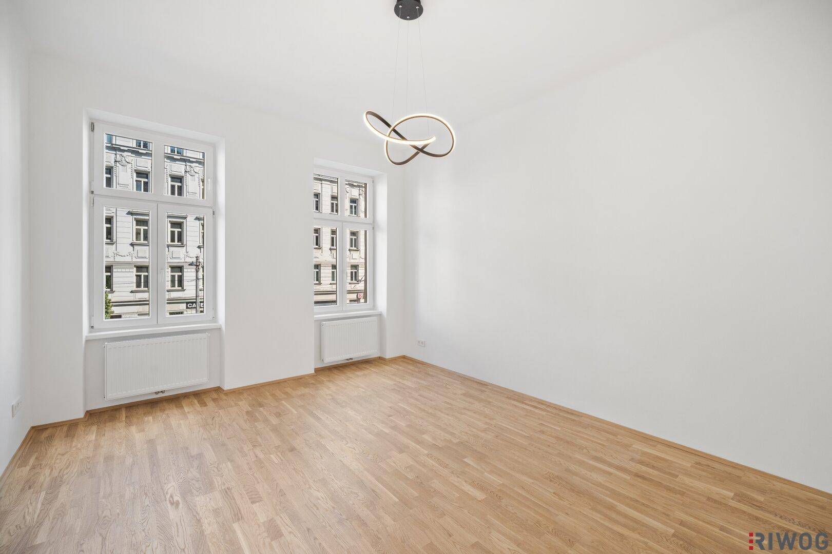 Leeres Zimmer mit weiß gestrichenen Wänden, Parkettboden und zwei Fenstern zur Straße.
