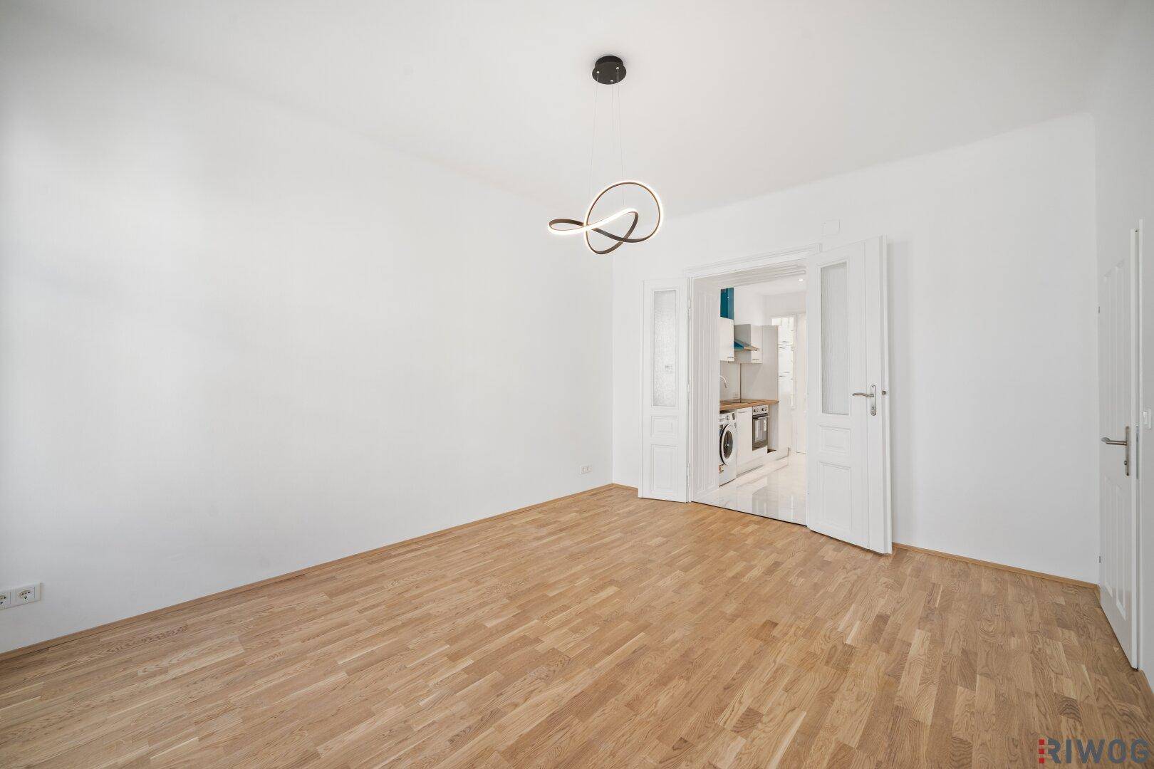 Geräumiges Zimmer mit Parkettboden und Zugang zu einer modernen Küche im Hintergrund.