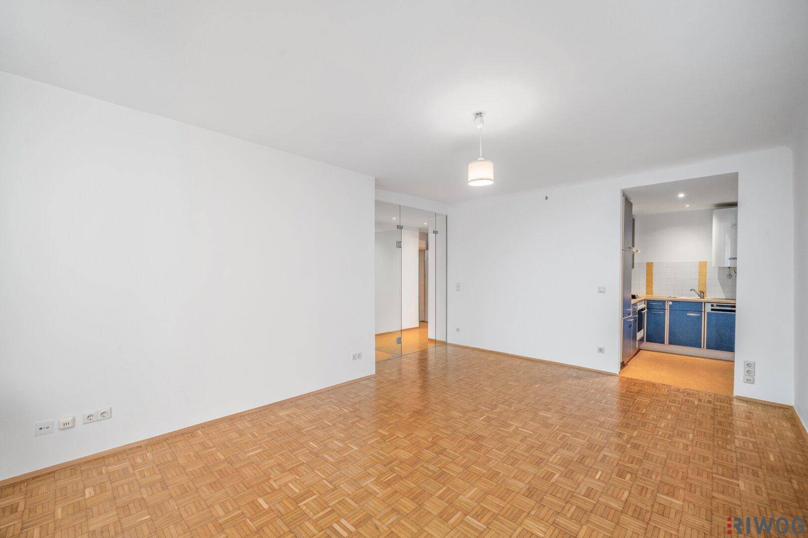 Geräumiges Zimmer mit Parkettboden, Glaswand und Zugang zu einer separaten Küche.