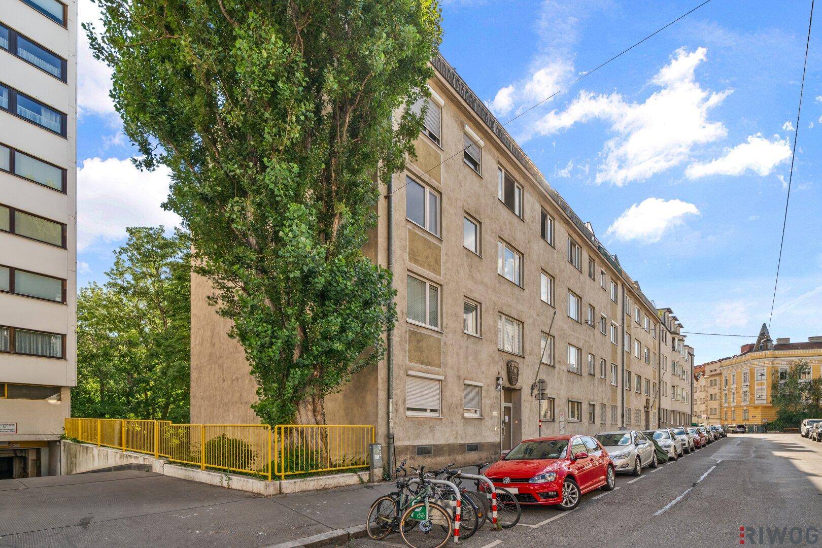 Mehrfamilienhaus in städtischer Umgebung mit Bäumen und parkenden Autos.