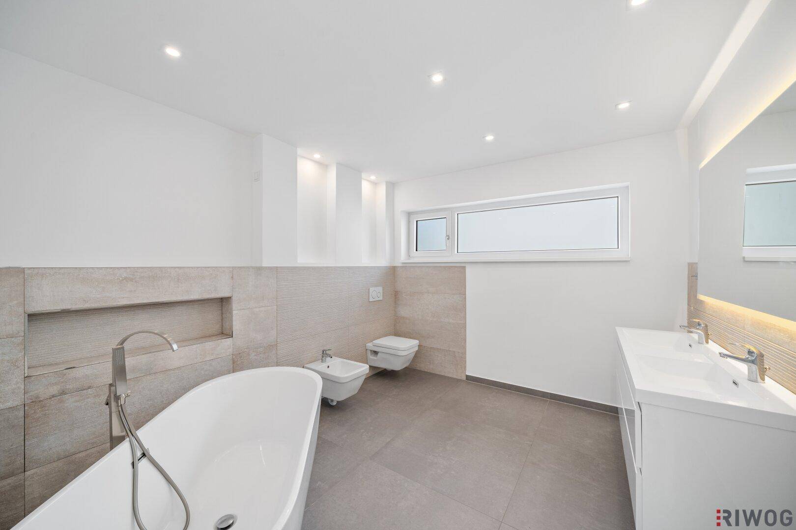 Helles Badezimmer mit freistehender Badewanne, wandhängendem WC, Bidet und Fenster.