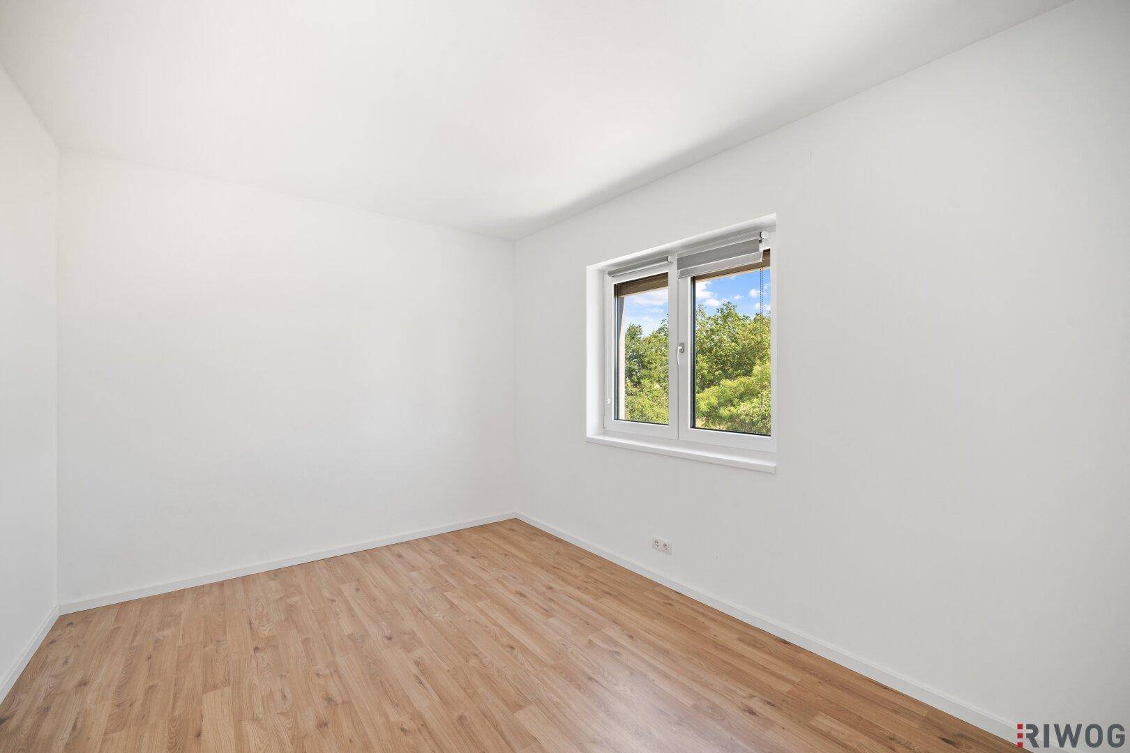 Helles Zimmer mit Holzboden und Fenster, das einen grünen Ausblick bietet.