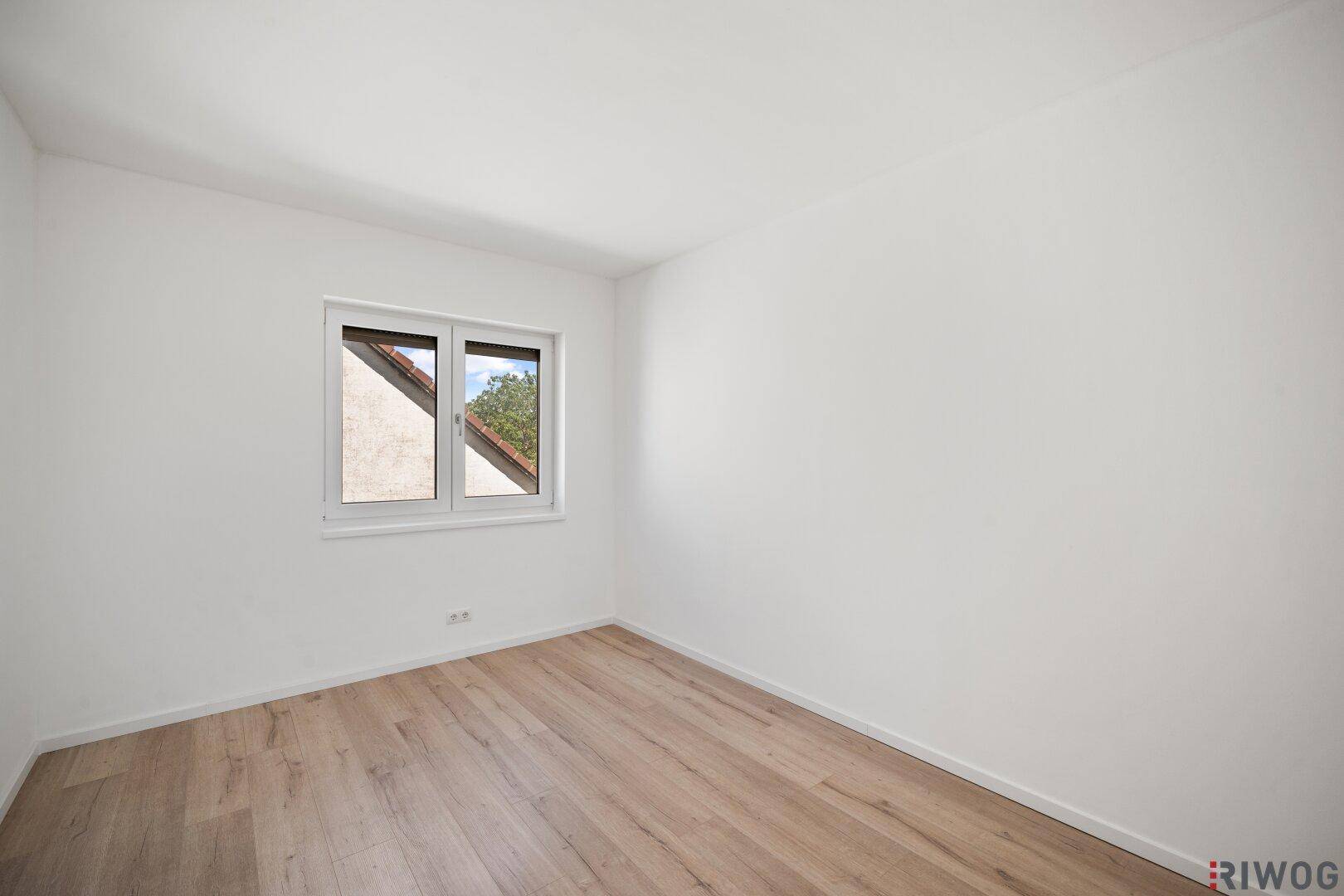 Helles Zimmer mit Holzboden und Fenster, das einen Blick auf die Dächer der Nachbarschaft bietet.