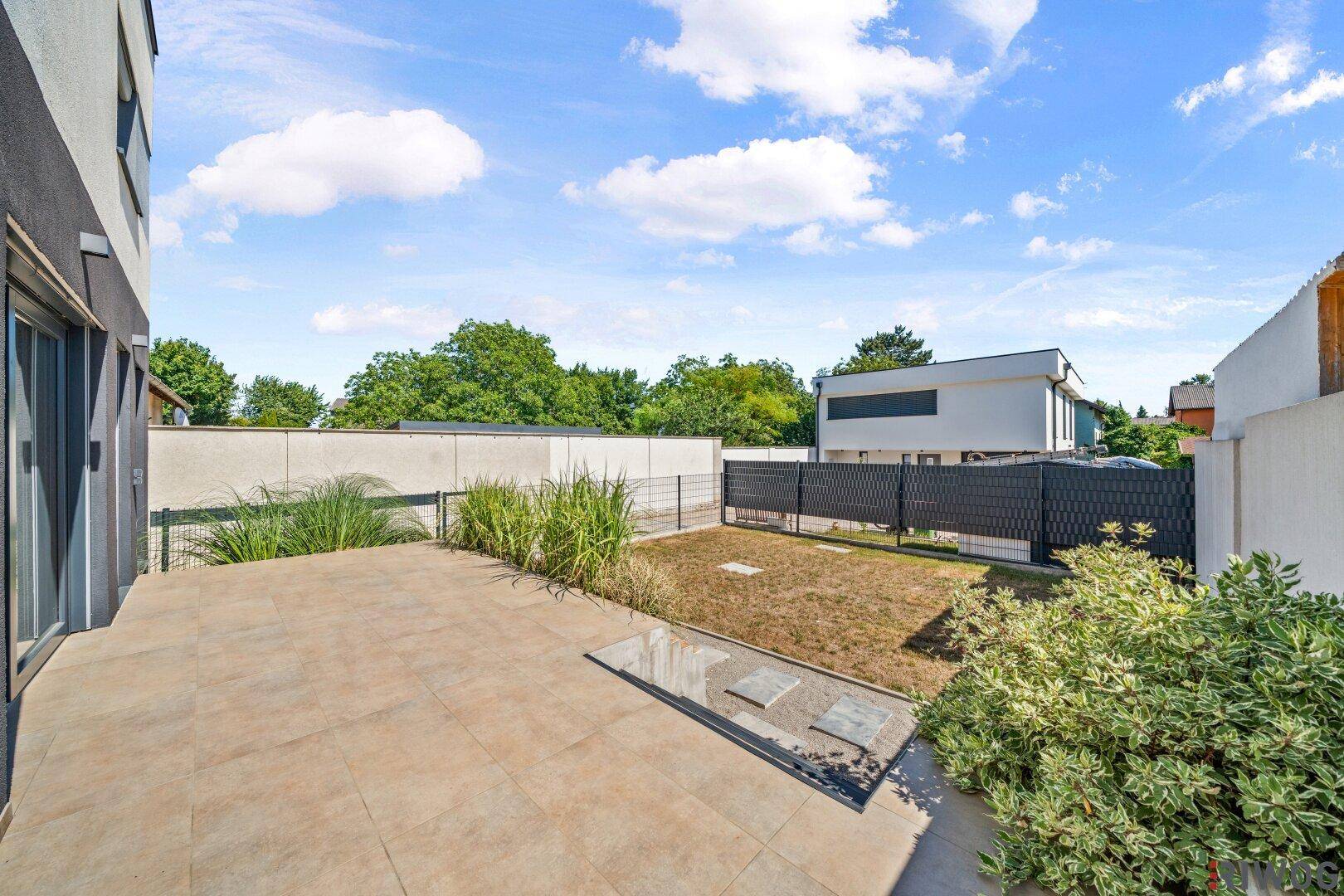 Heller Terrassenbereich mit beigen Fliesen und angrenzendem Garten, ideal für Entspannung im Freien.