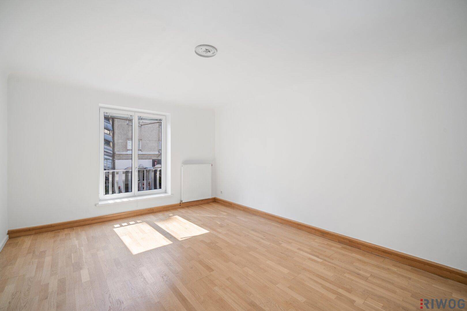 Helles Zimmer mit Parkettboden und einem geöffneten Fenster, das einen französischen Balkon andeutet.