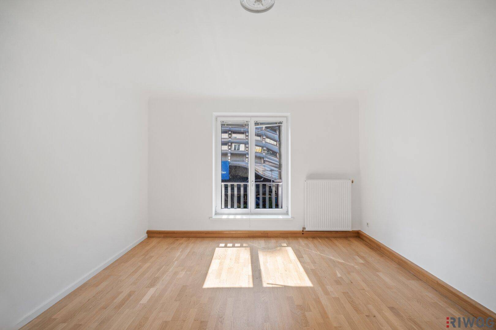 Freundliches Zimmer mit Parkettboden, weißen Wänden und einem Fenster mit Blick auf die Umgebung.