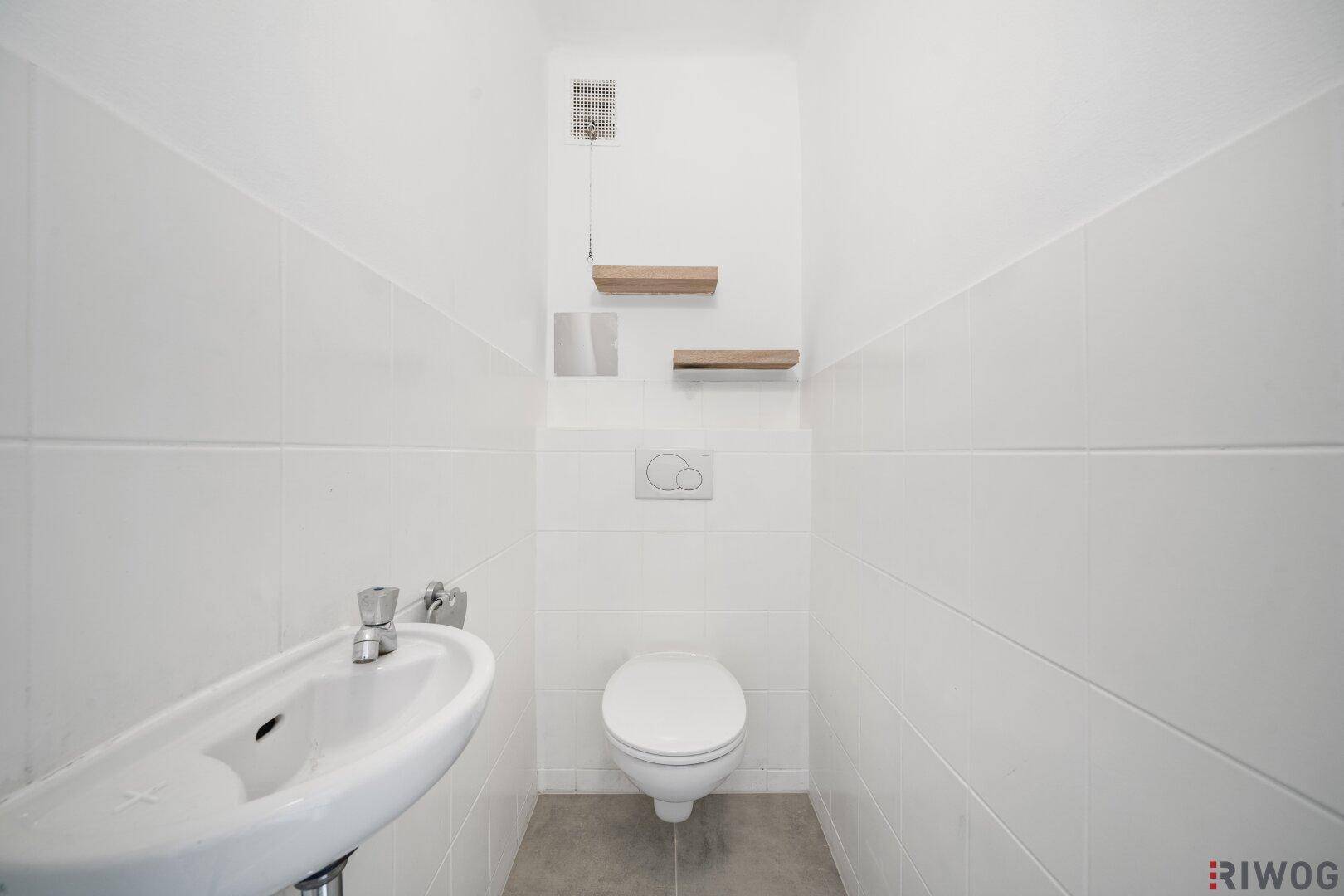 Separates WC mit wandhängender Toilette, kleinem Waschbecken und weißen Fliesen.