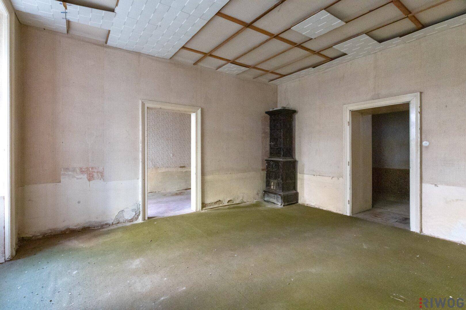 Sanierungsbedürftiger Raum mit traditionellem Kachelofen und beschädigter Decke.