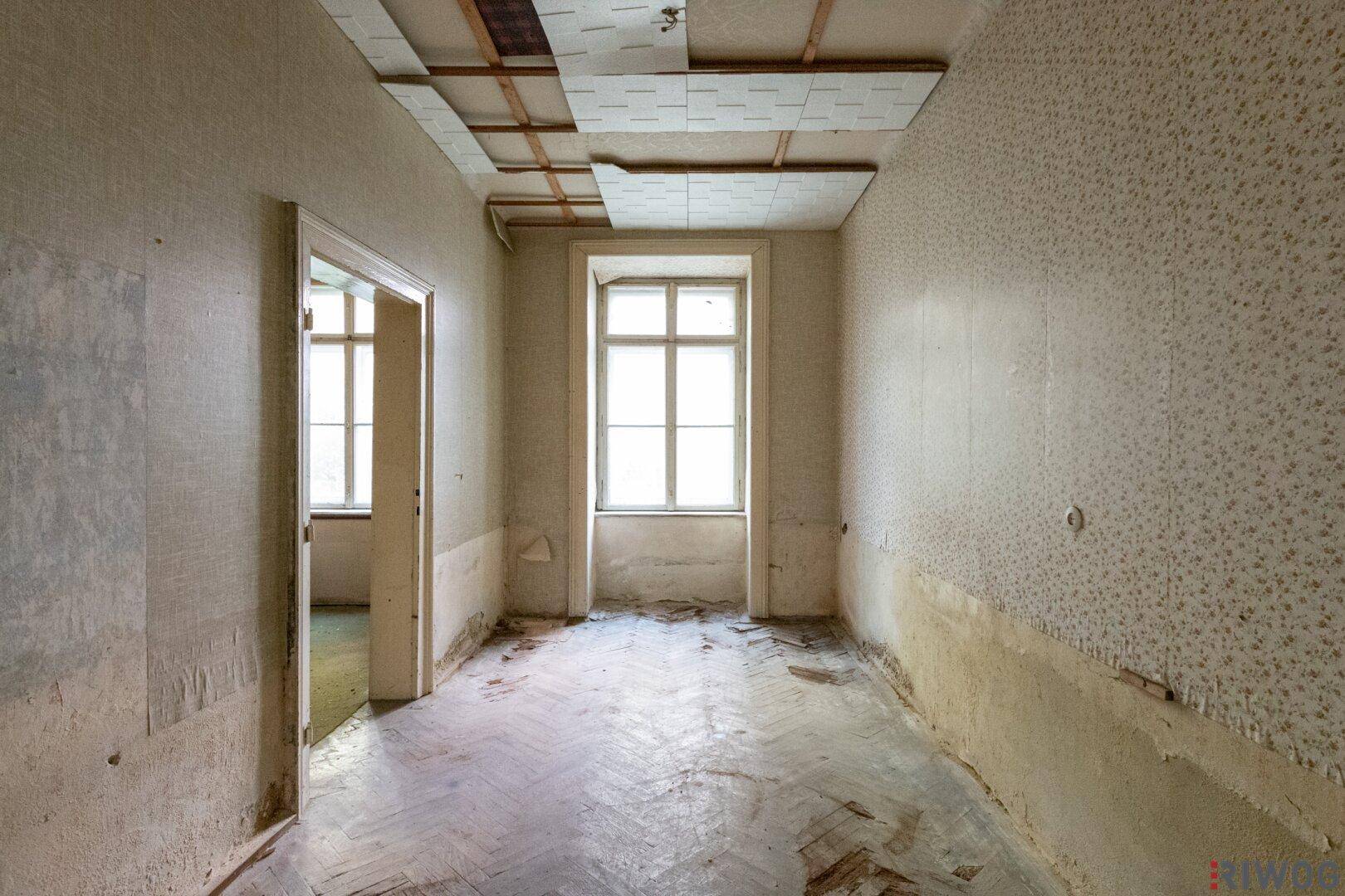 Unmöblierter Raum mit beschädigtem Parkettboden und alten Tapetenresten.