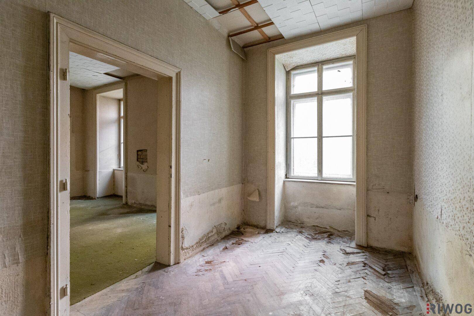 Leerer Raum mit altem Fischgrätparkett und renovierungsbedürftigen Wänden.