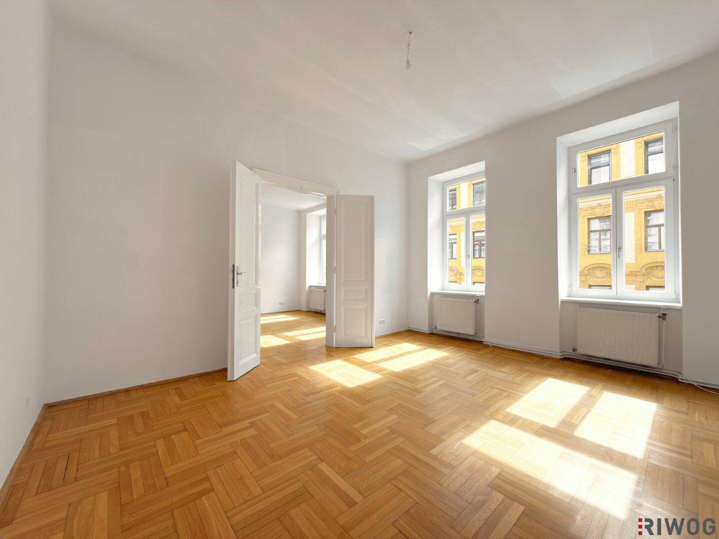Geräumiges, leeres Zimmer mit Fischgrätparkett und hohen Decken, viel Tageslicht.