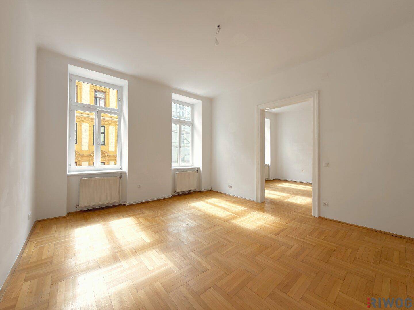 Geräumiges, leeres Zimmer mit Fischgrätparkett und hohen Decken, viel Tageslicht.