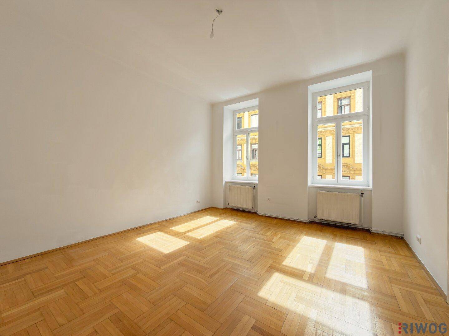 Helles, leeres Zimmer mit Parkettboden und zwei großen Fenstern, lichtdurchflutet.