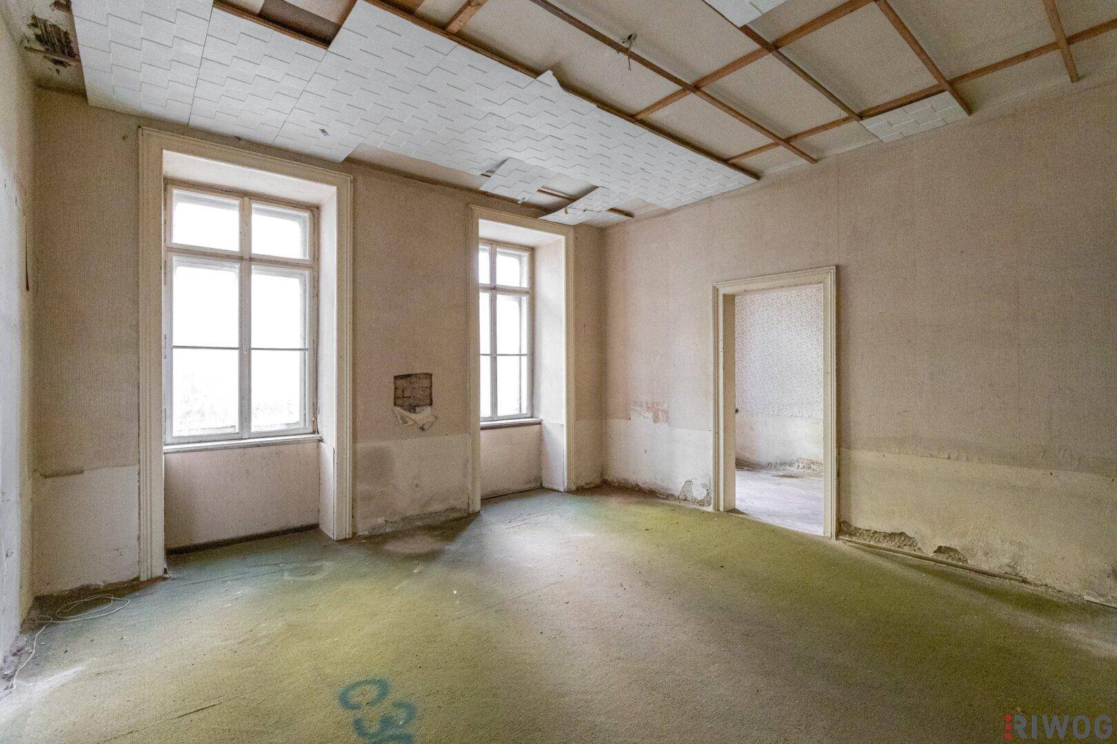 Renovierungsbedürftiger Raum mit alten Fenstern und beschädigter Deckenverkleidung.