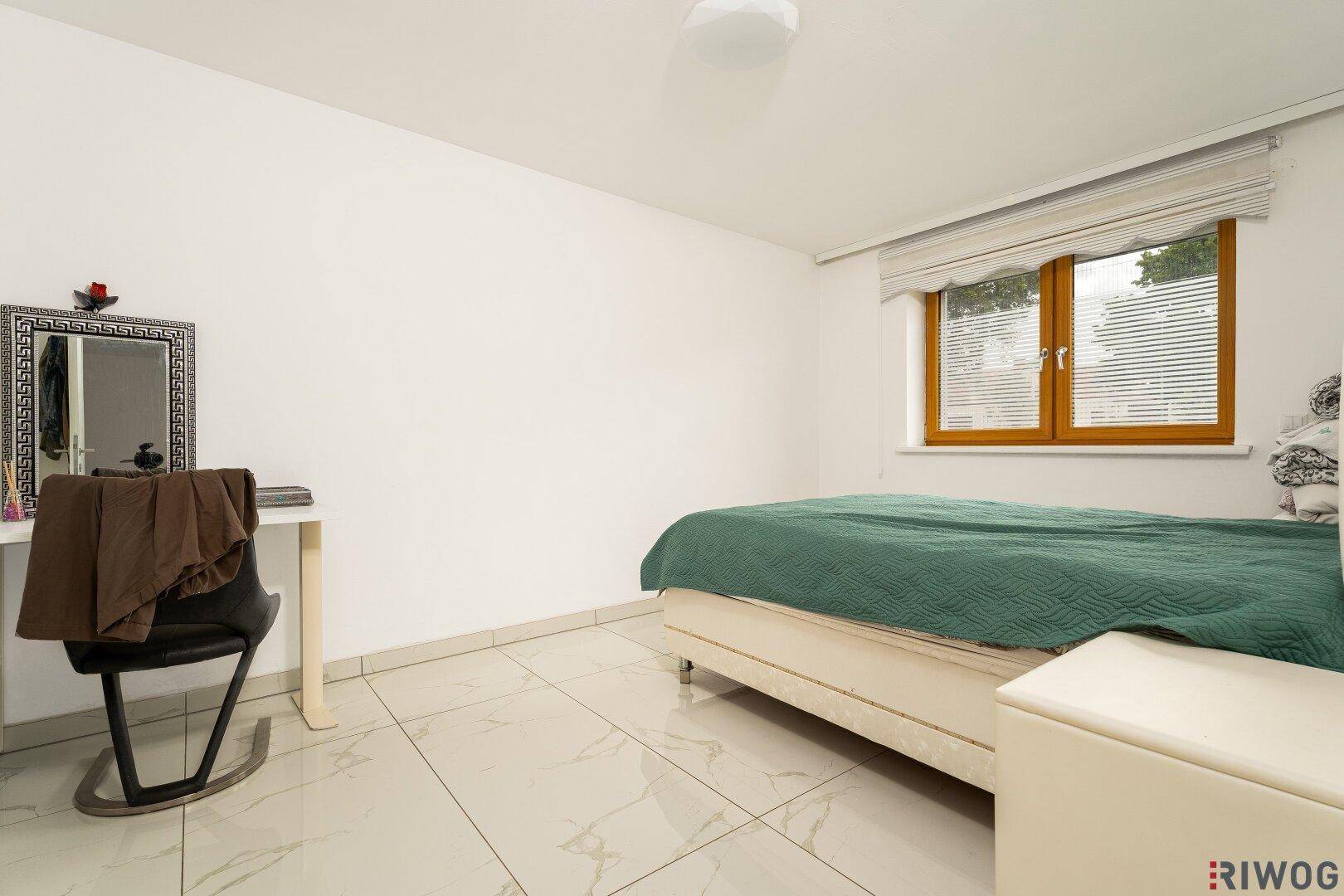 Schlafzimmer mit hellen Fliesen, einem Bett mit grüner Decke und einem Schminktisch am Fenster.