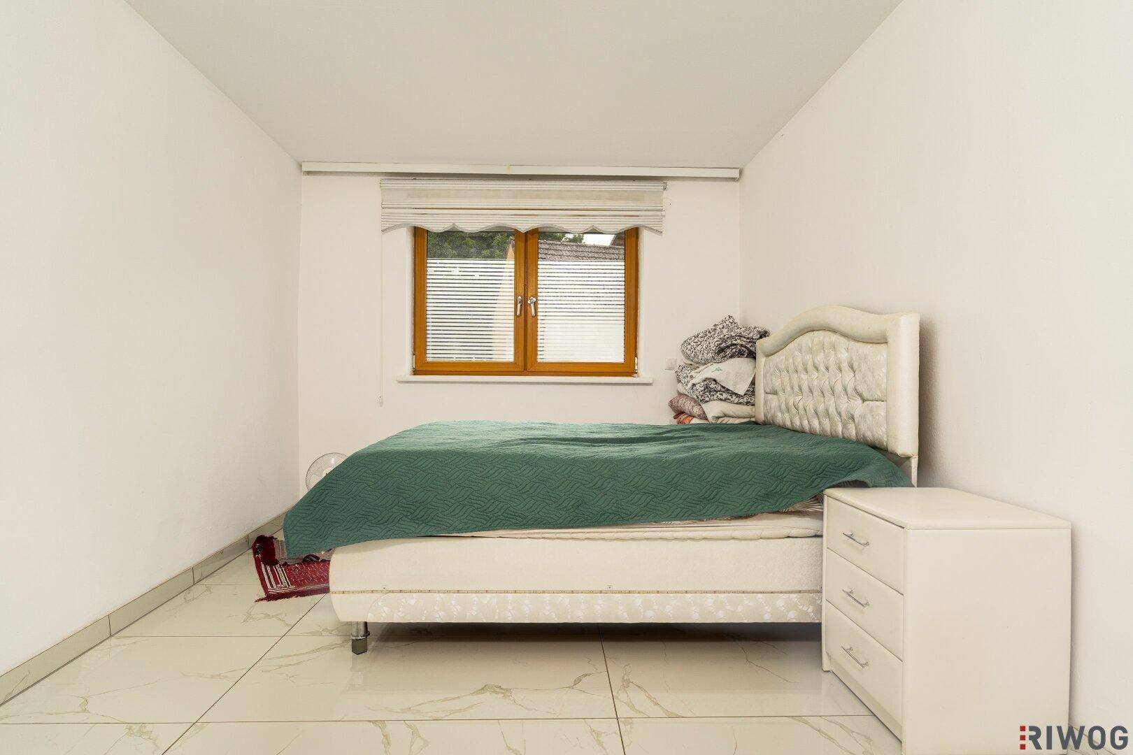 Schlafzimmer mit modernem Bett, weißem Nachttisch und Fenster mit Jalousien.