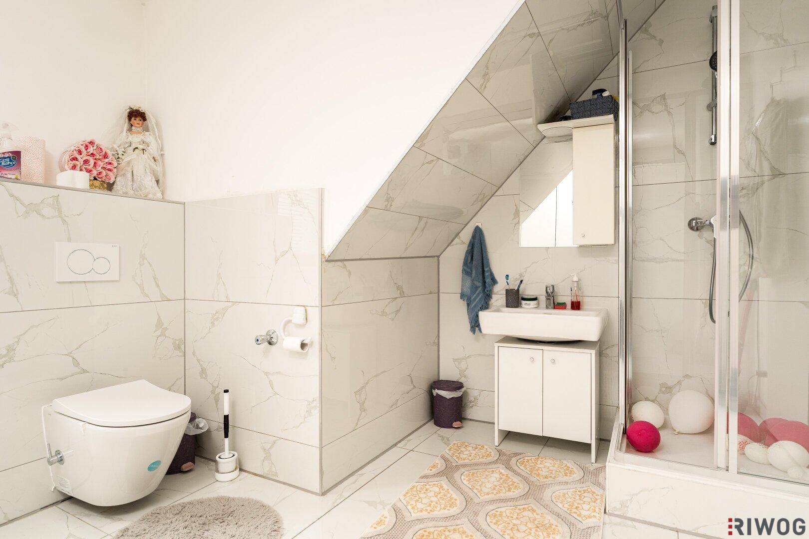 Modernes Badezimmer mit Marmoroptik-Fliesen, Toilette, Waschbecken und Duschkabine unter Dachschräge.