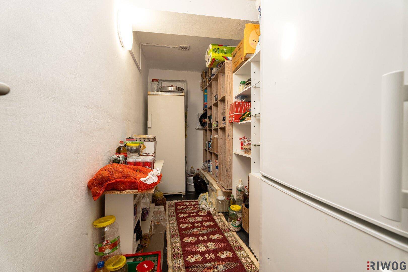 Schmaler Vorratsraum mit Regalen, einem Kühlschrank und zahlreichen gelagerten Lebensmitteln und Haushaltsartikeln.