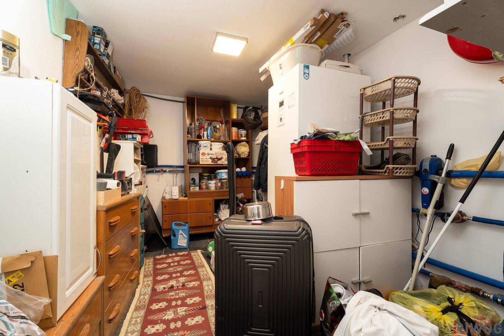 Unaufgeräumter Abstellraum mit Regalen, Kommoden und einem Kühlschrank, der diverse Haushaltsgegenstände beherbergt.