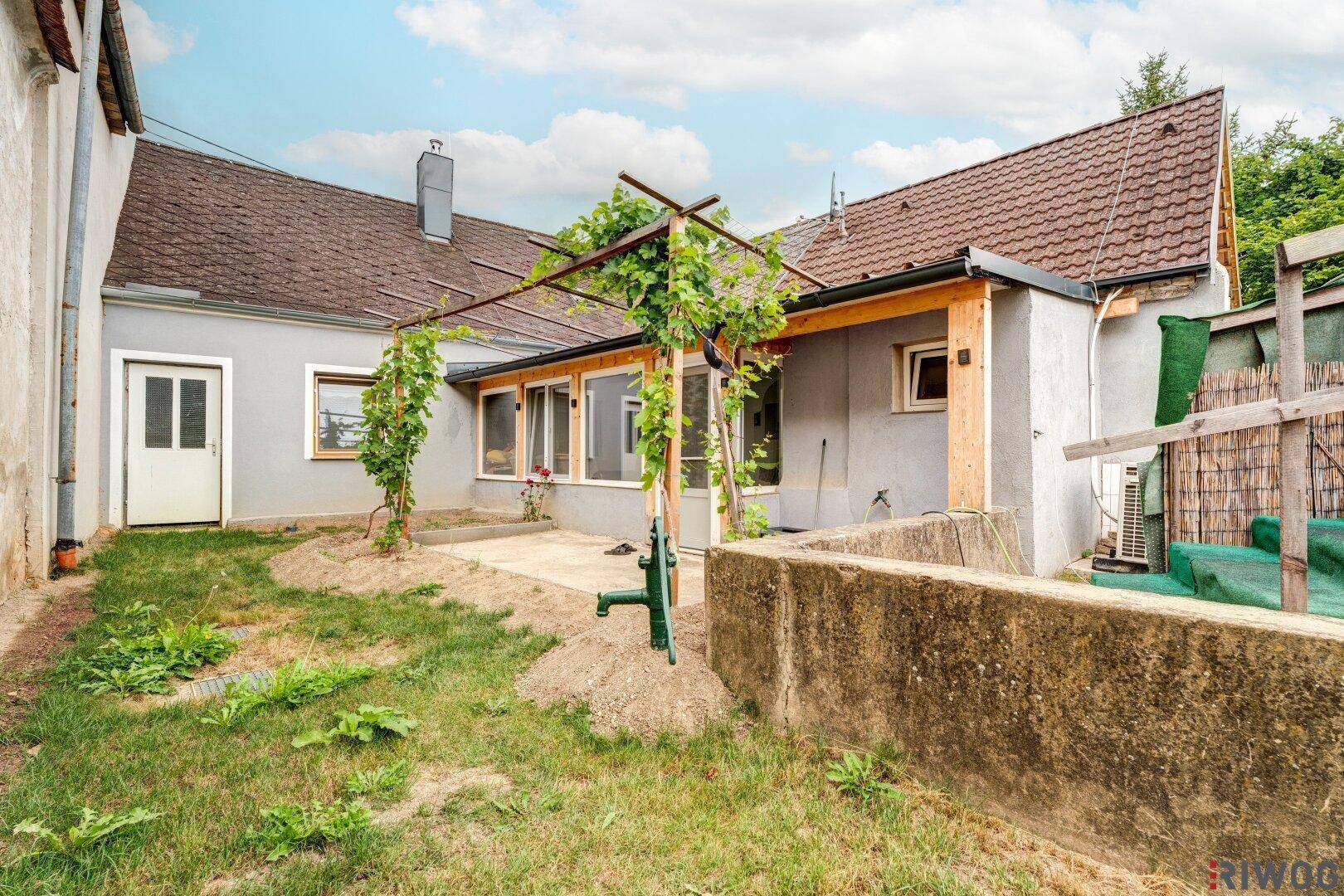 Hinterhof mit kleiner Terrasse, Wasserpumpe und einem Anbau, umgeben von Grünflächen.