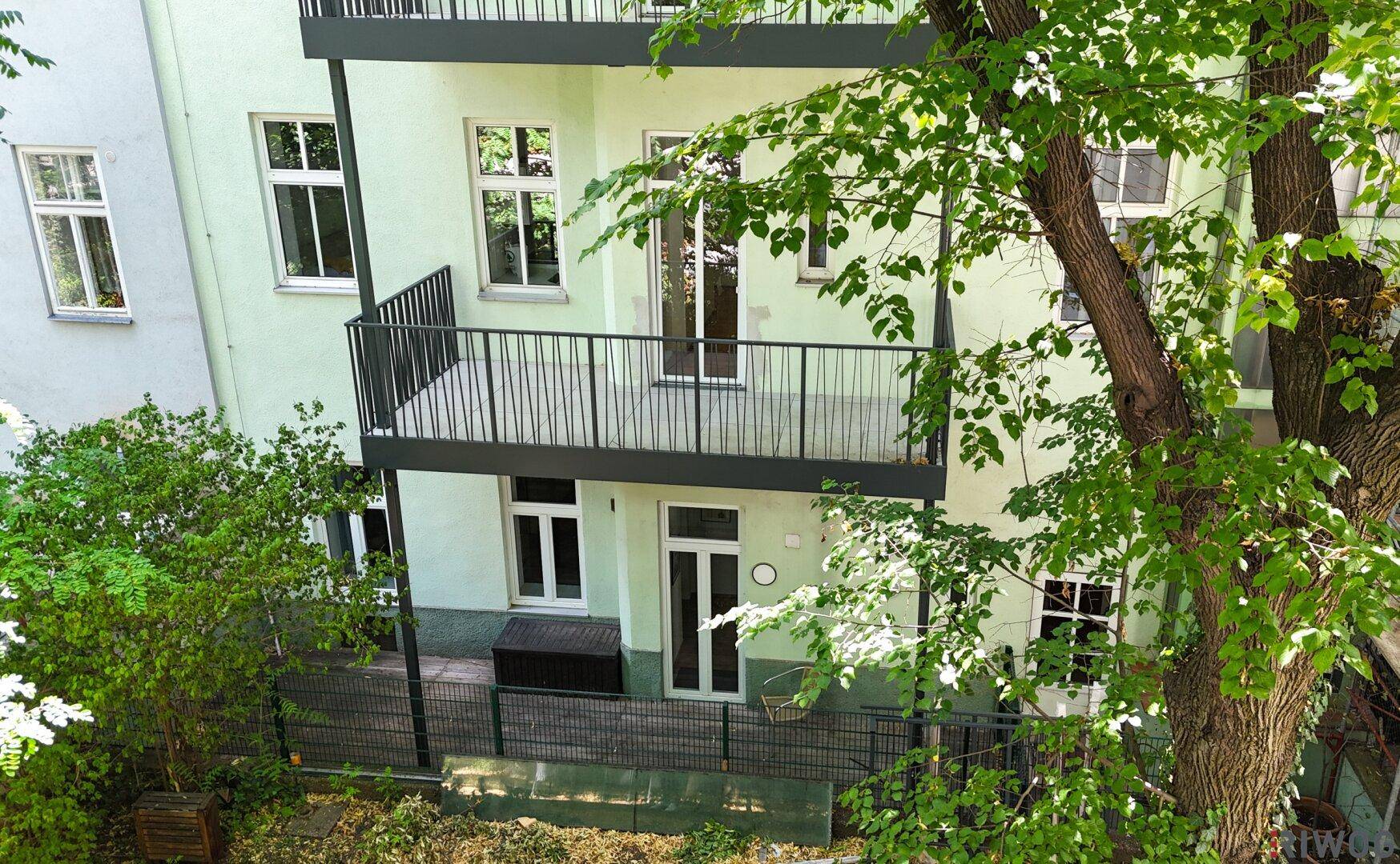 Unterer Bereich einer Gebäudefassade mit Balkon und Zugang zu einem begrünten Außenbereich.