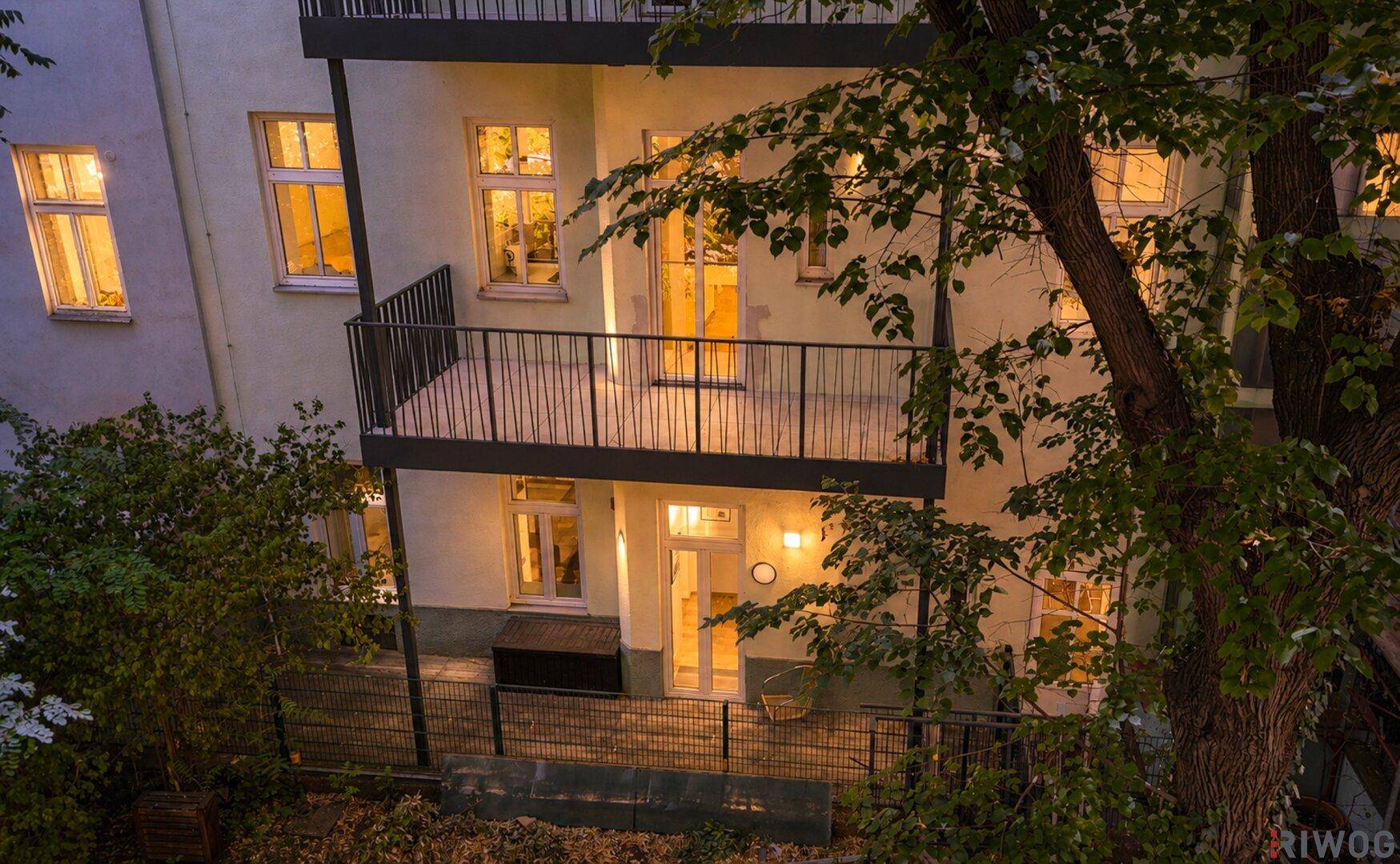Rückansicht des Gebäudes bei Nacht mit beleuchteten Balkonen und einer Terrasse, umgeben von Bäumen.