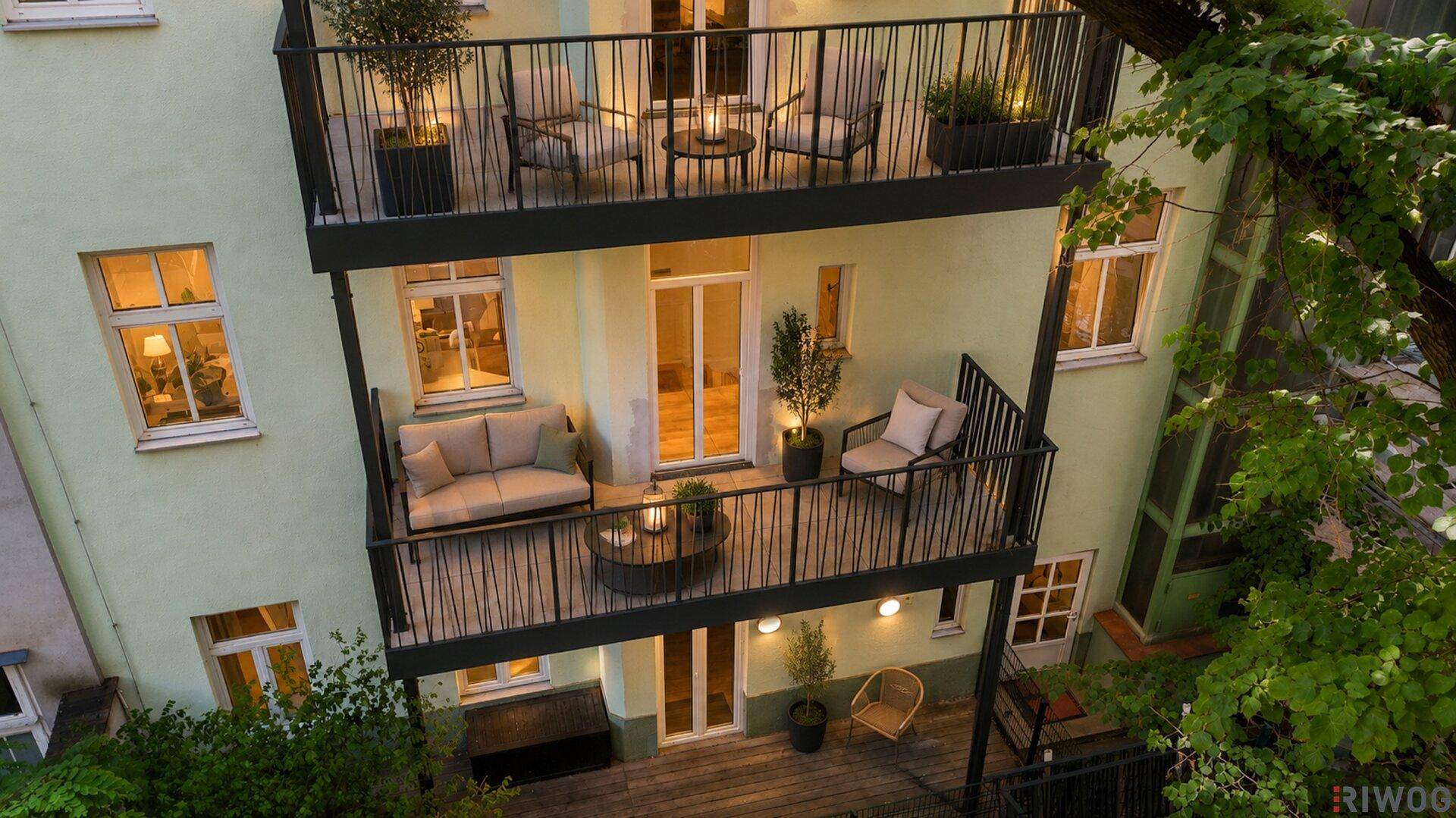 Helle Rückansicht des Gebäudes mit möblierten Balkonen und einer Terrasse, umgeben von Bäumen.