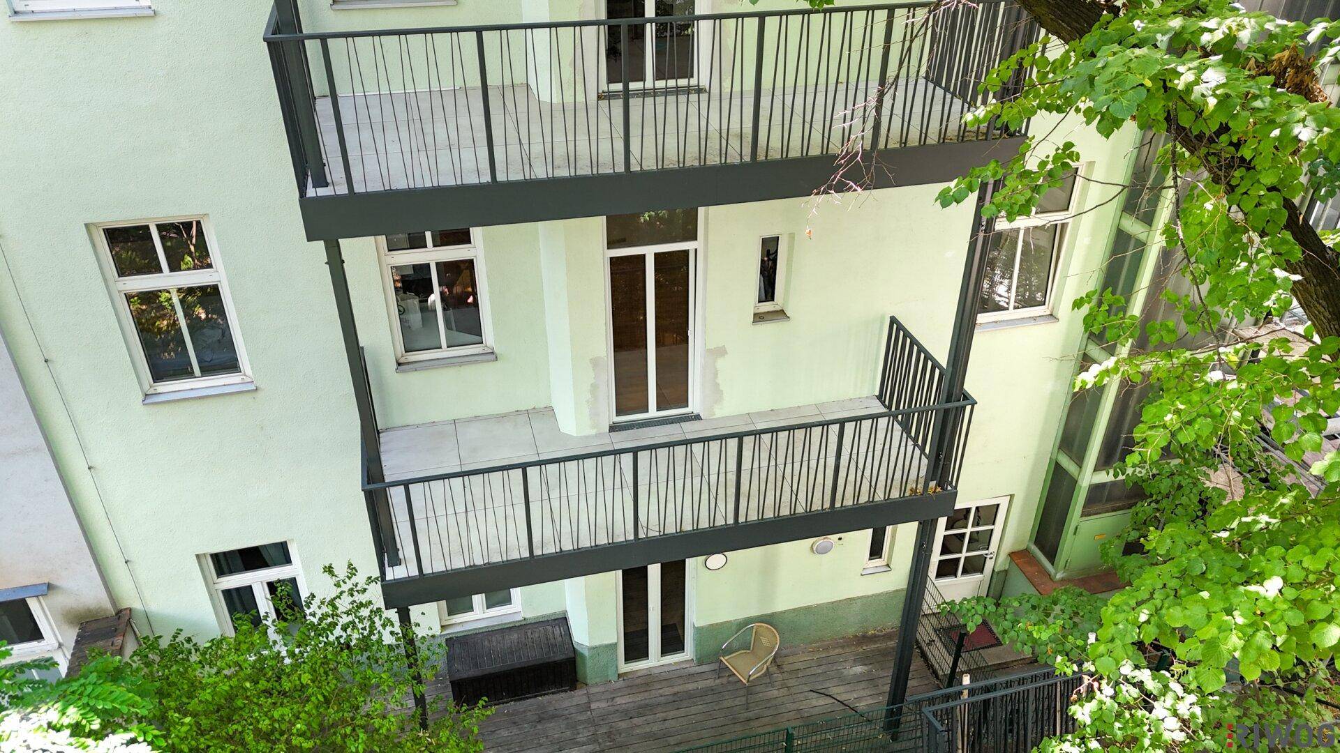 Helle Rückansicht des Gebäudes am Tag mit mehreren Balkonen und einer Terrasse, umgeben von viel Grün.