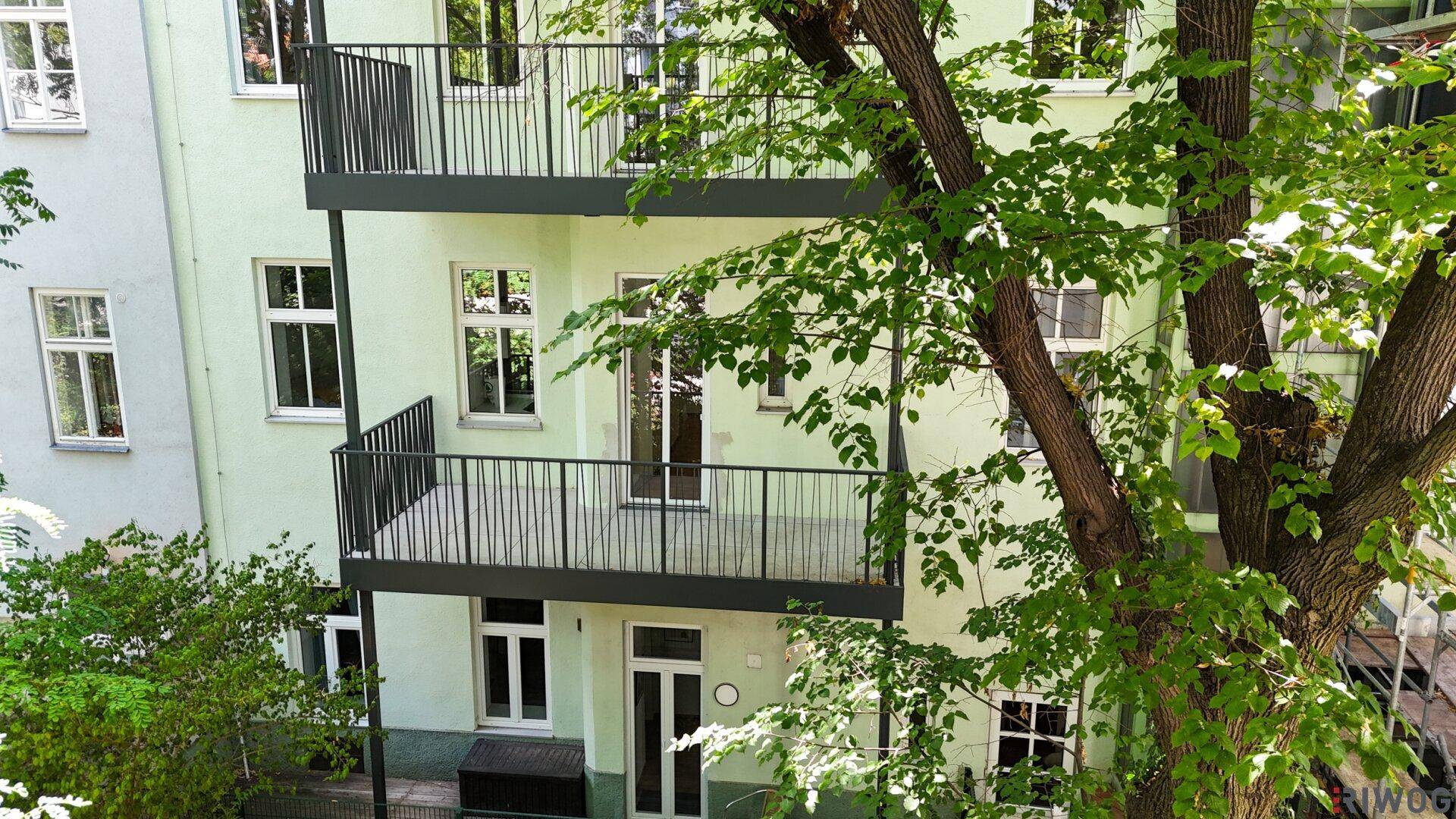 Helle Fassade mit zwei leeren Balkonen und umgebenden Bäumen im Tageslicht.