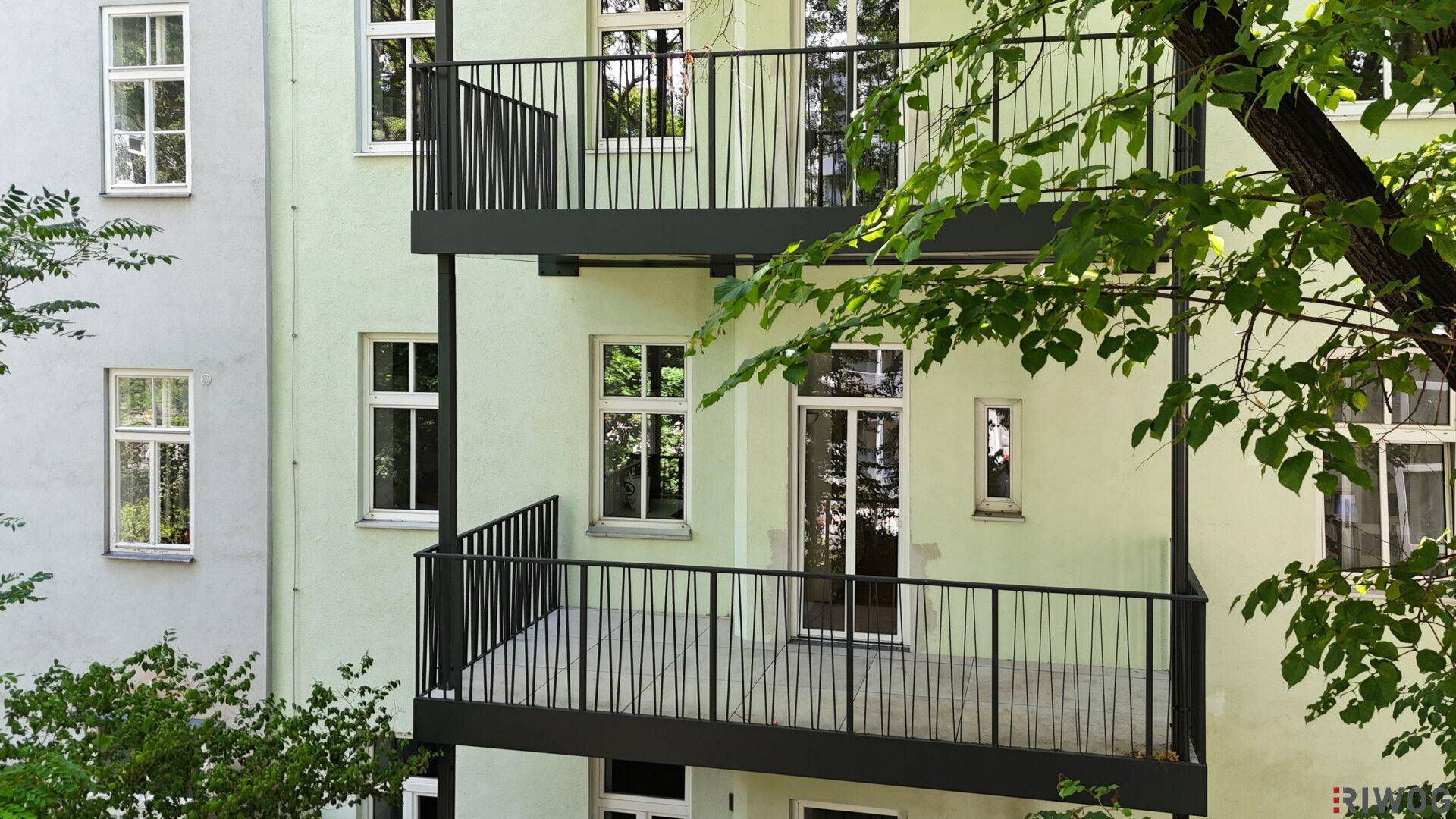 Tageslichtaufnahme der Gebäudefassade mit zwei leeren Balkonen und grüner Umgebung.