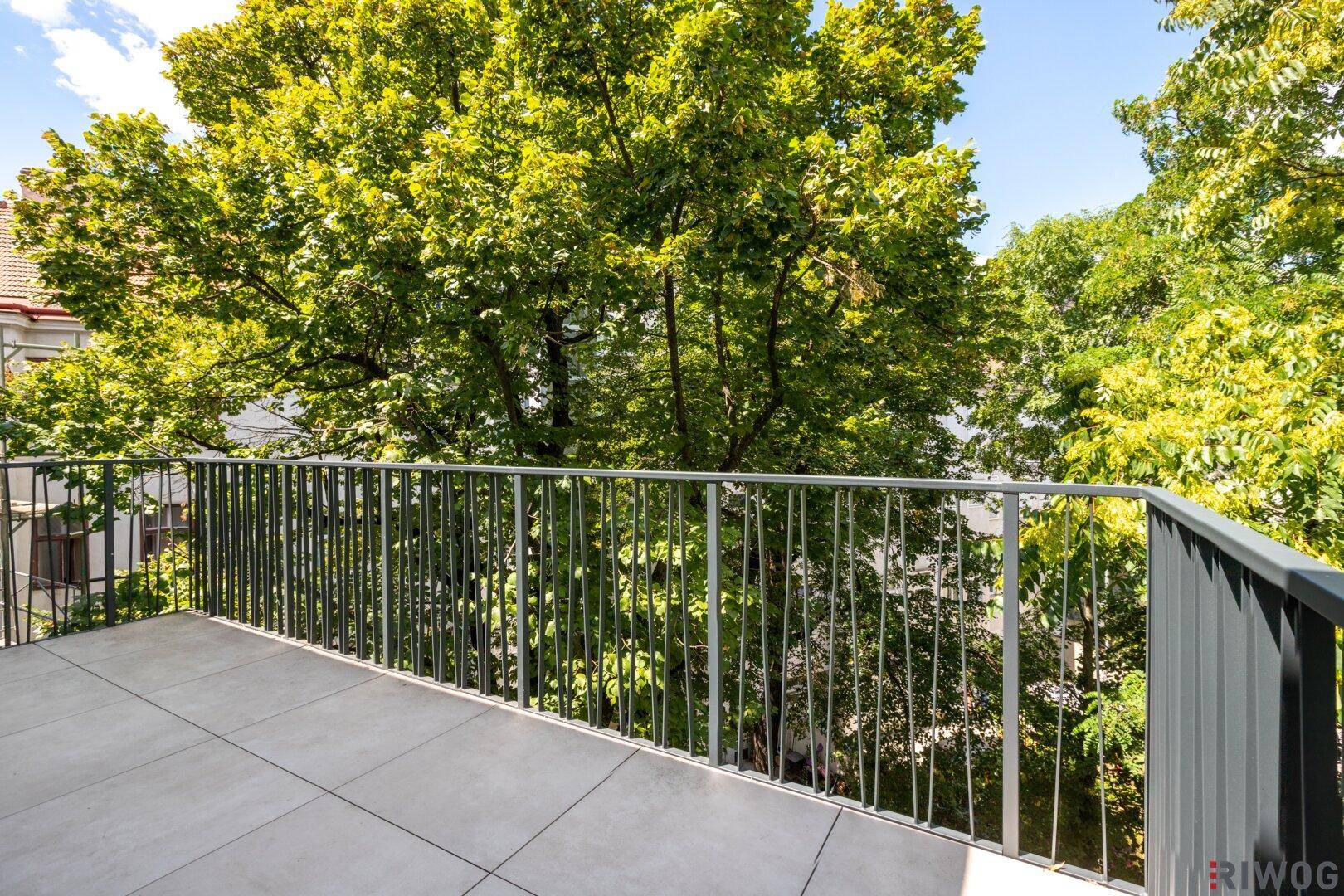 Geräumiger Balkon mit Fliesenboden und Metallgeländer, umgeben von üppigem Grün an einem hellen Tag.