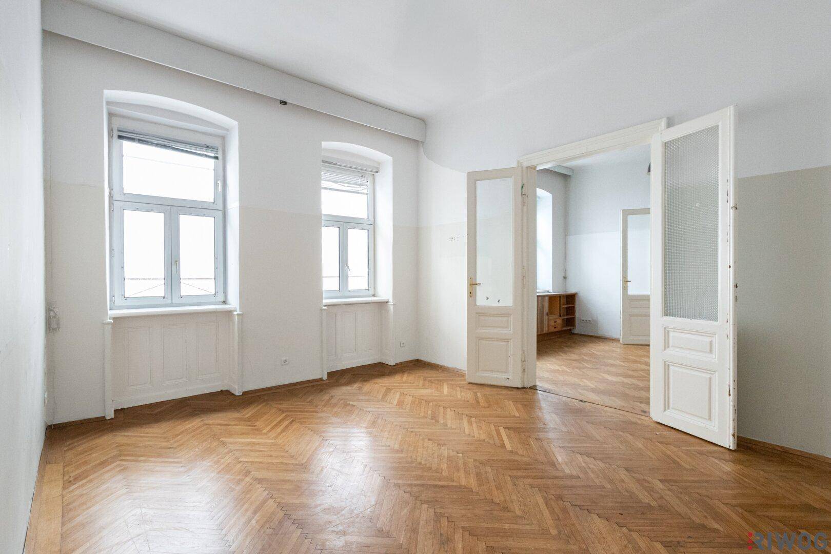 Geräumiges Zimmer mit Fischgrätparkett, hohen Decken und großen Fenstern, die viel Licht hereinlassen.