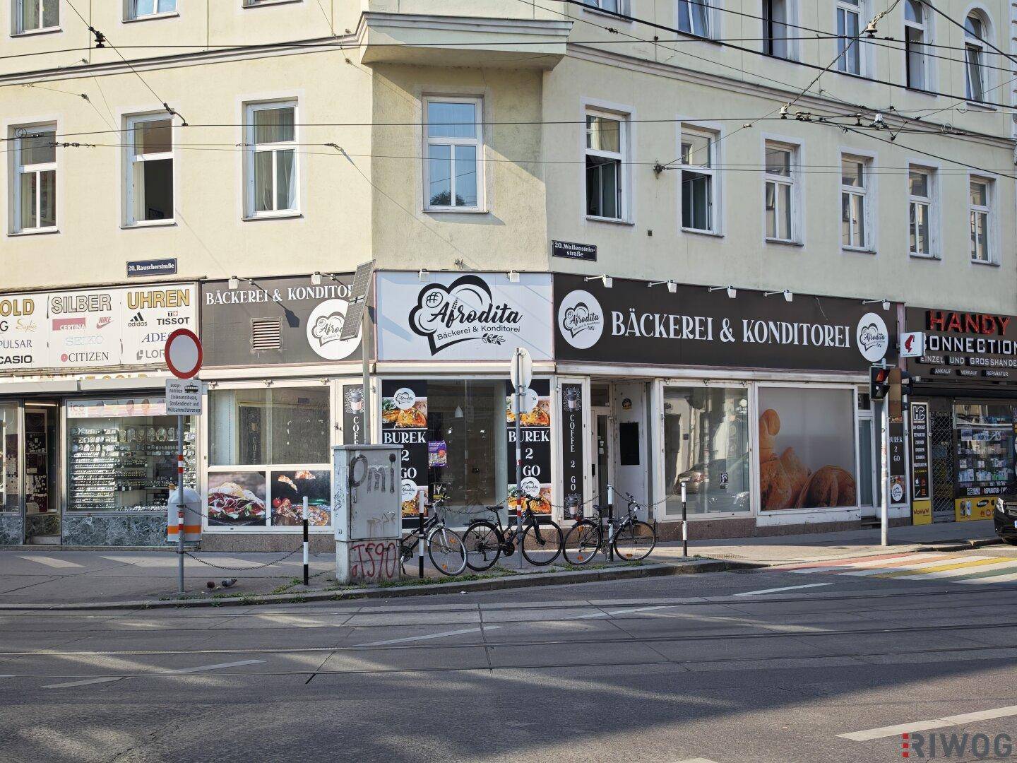 Nahaufnahme der Ladenfronten im Erdgeschoss eines Eckgebäudes mit Bäckereien und anderen Geschäften.