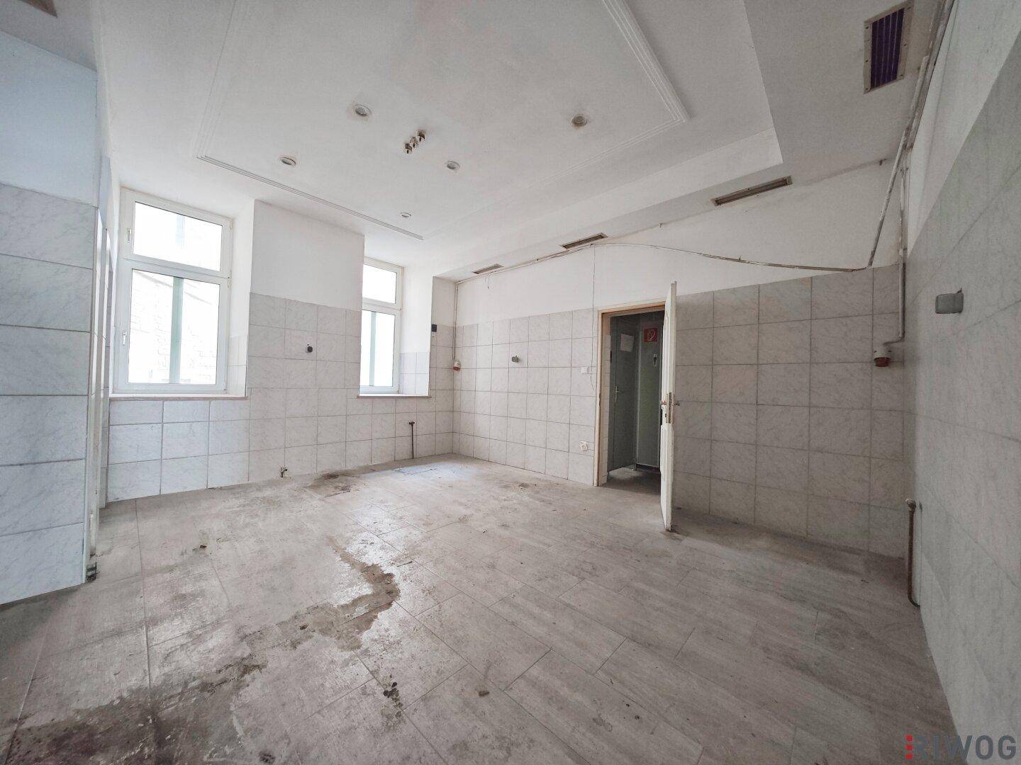 Großer, gefliester Raum mit zwei Fenstern, der viel Tageslicht bietet und renovierungsbedürftig ist.