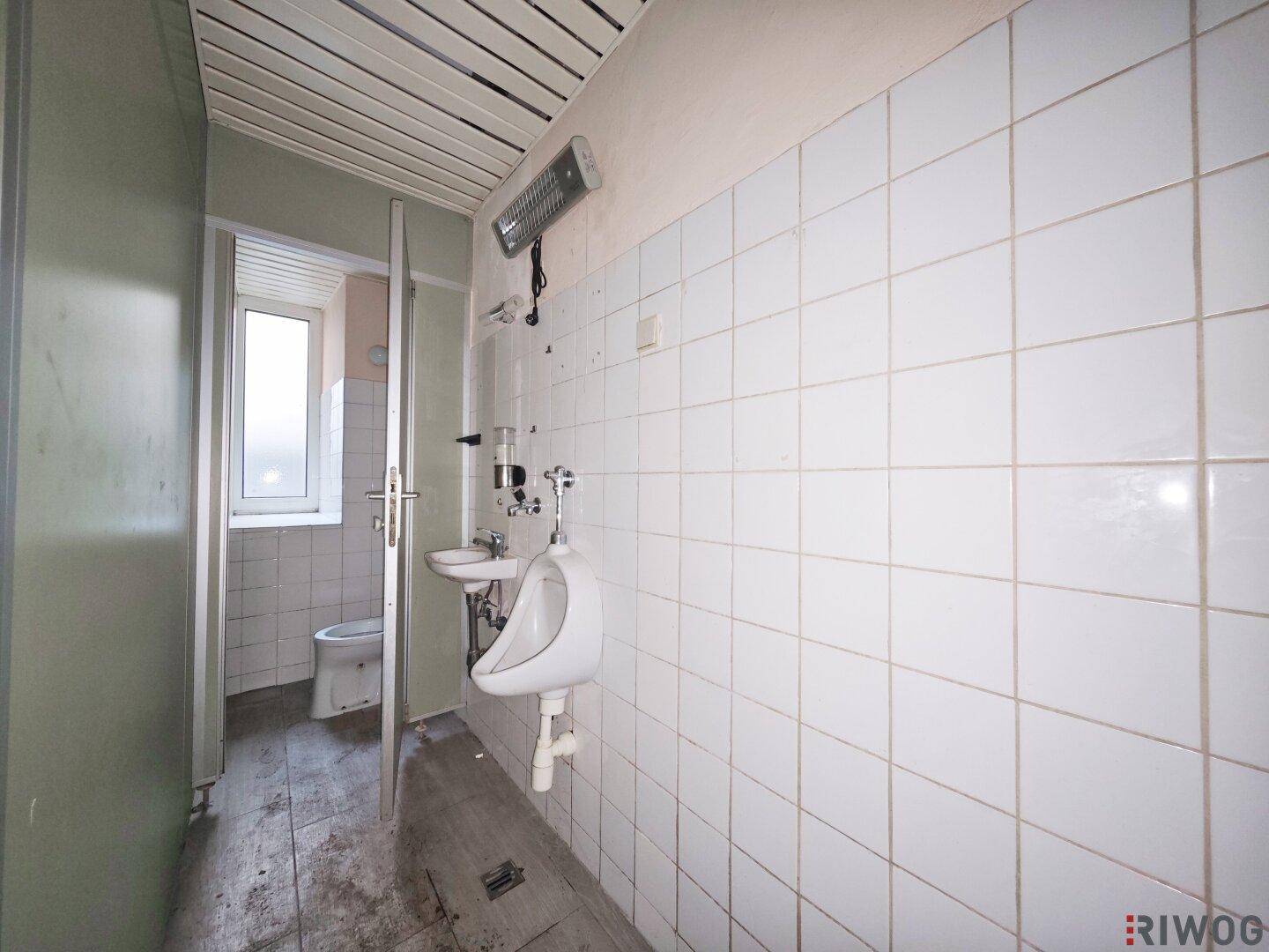 Gefliester Toilettenraum mit Urinal und separater WC-Kabine, renovierungsbedürftig.