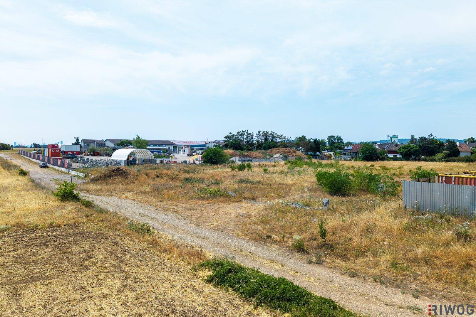 Weitläufiges, trockenes Feld mit einem Feldweg und industriellen Gebäuden im Hintergrund unter blauem Himmel.