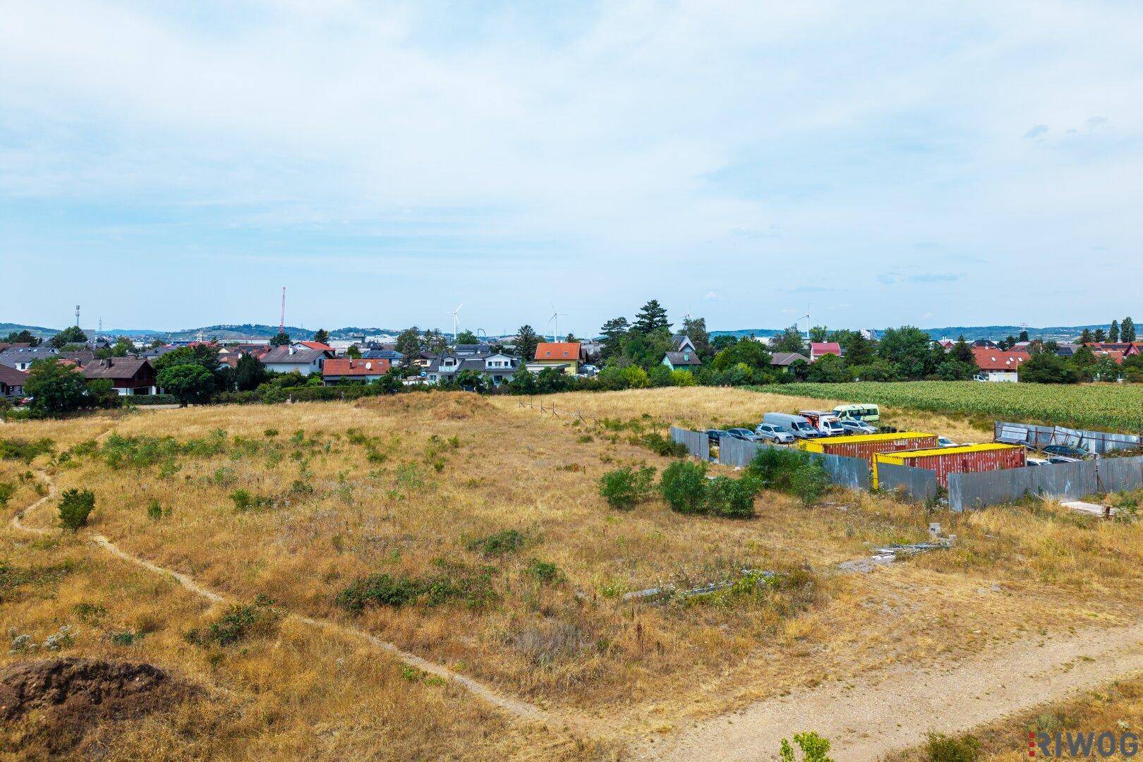 Weitläufiges, trockenes Feld mit temporären Strukturen und Fahrzeugen am Rand, umgeben von ländlicher Bebauung.
