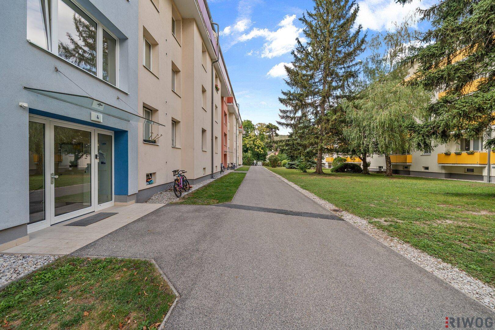 Eingangsbereich eines Mehrfamilienhauses mit gepflegter Außenanlage und Bäumen.