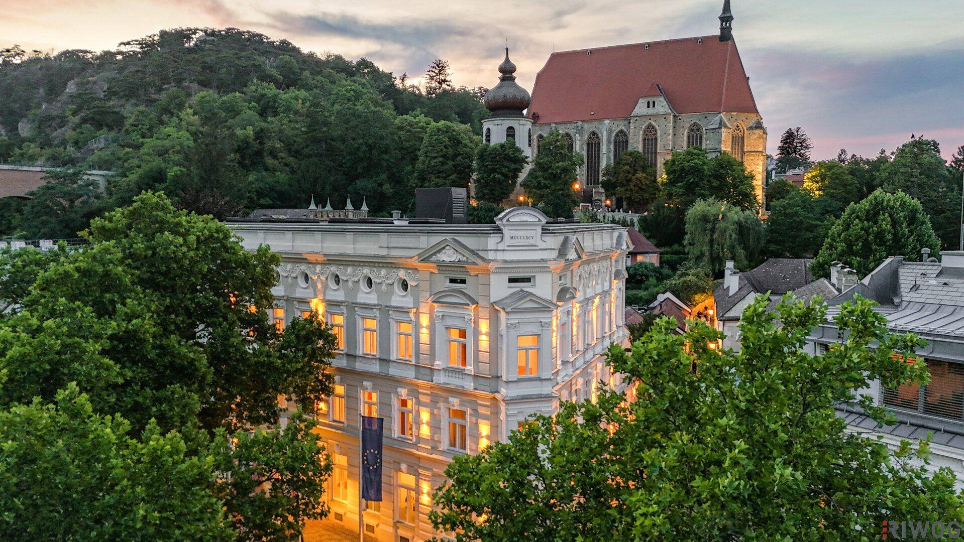 Historisches Gebäude mit Blick auf eine Kirche und grüne Hügel bei Sonnenuntergang.