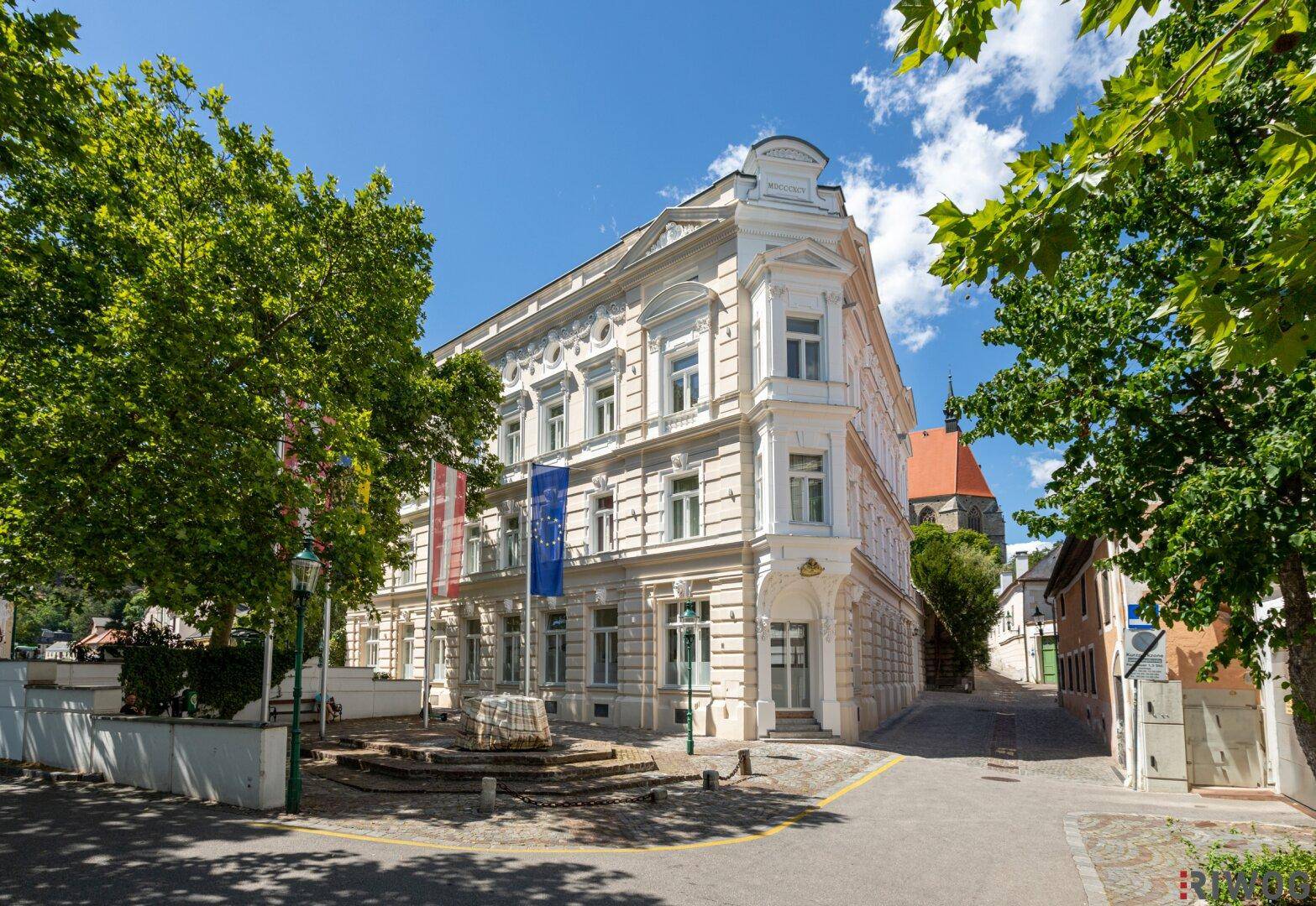 Historisches Gebäude mit eleganter Fassade, umgeben von Bäumen und einer ruhigen Straße an einem sonnigen Tag.