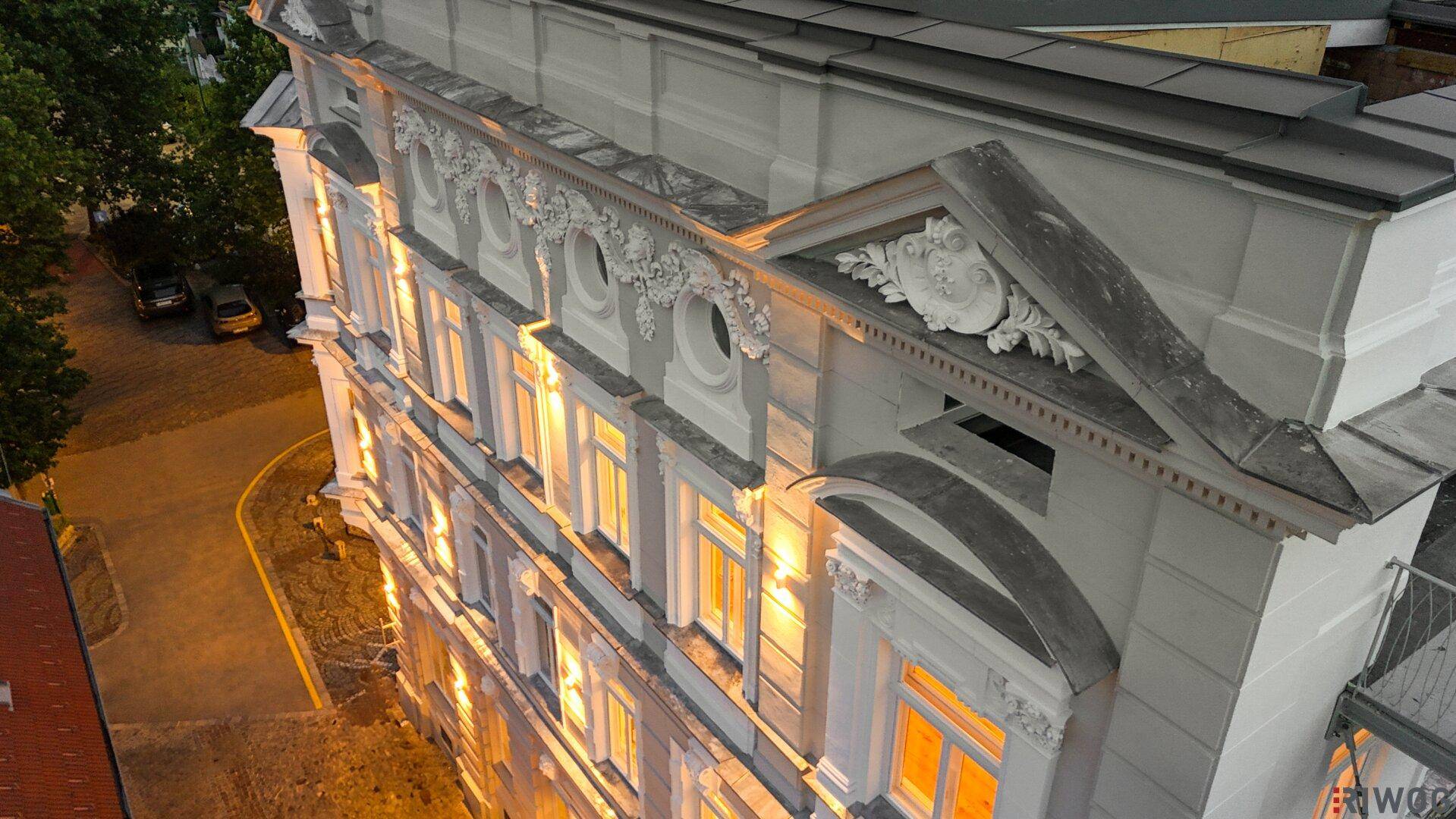 Detailaufnahme der historischen Fassade mit kunstvollen Verzierungen und beleuchteten Fenstern bei Dämmerung.