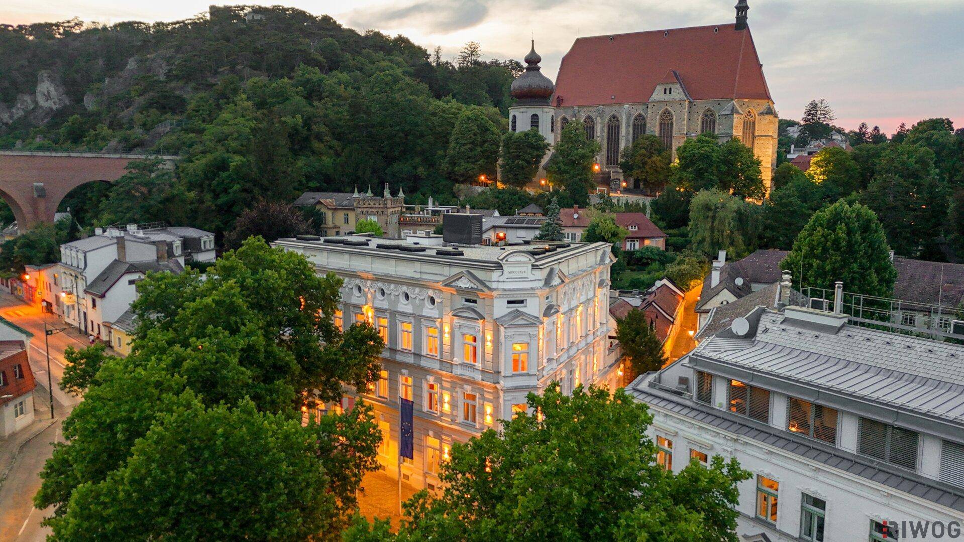 Historisches Gebäude mit beleuchteter Fassade und Blick auf eine Kirche und grüne Hügel bei Dämmerung.